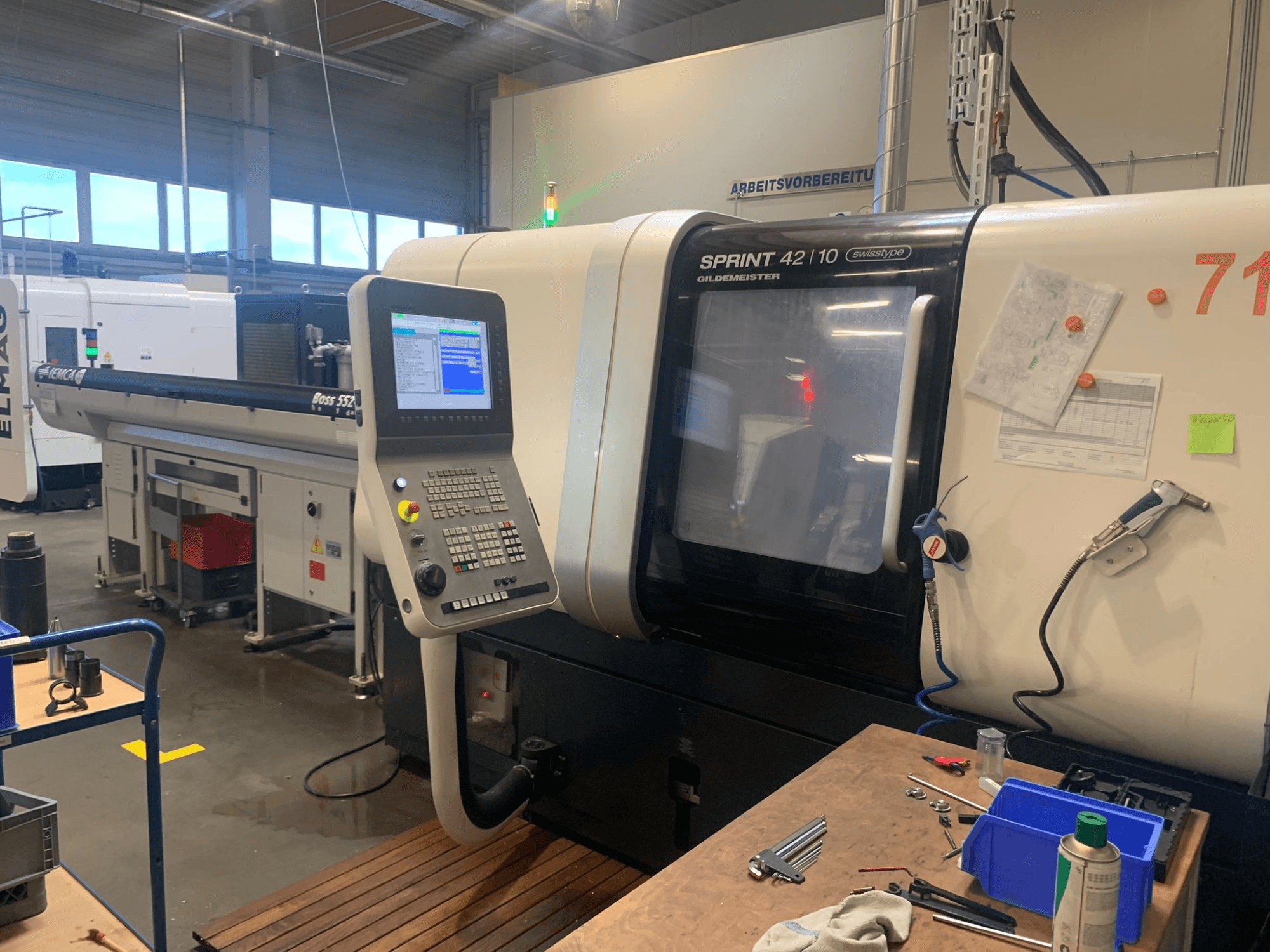 DMG MORI Sprint 42/10 linijinės šveicariško tipo tekinimo staklės, vaizdas iš priekio, su valdymo skydeliu ir darbo vietos netvarkingumu.