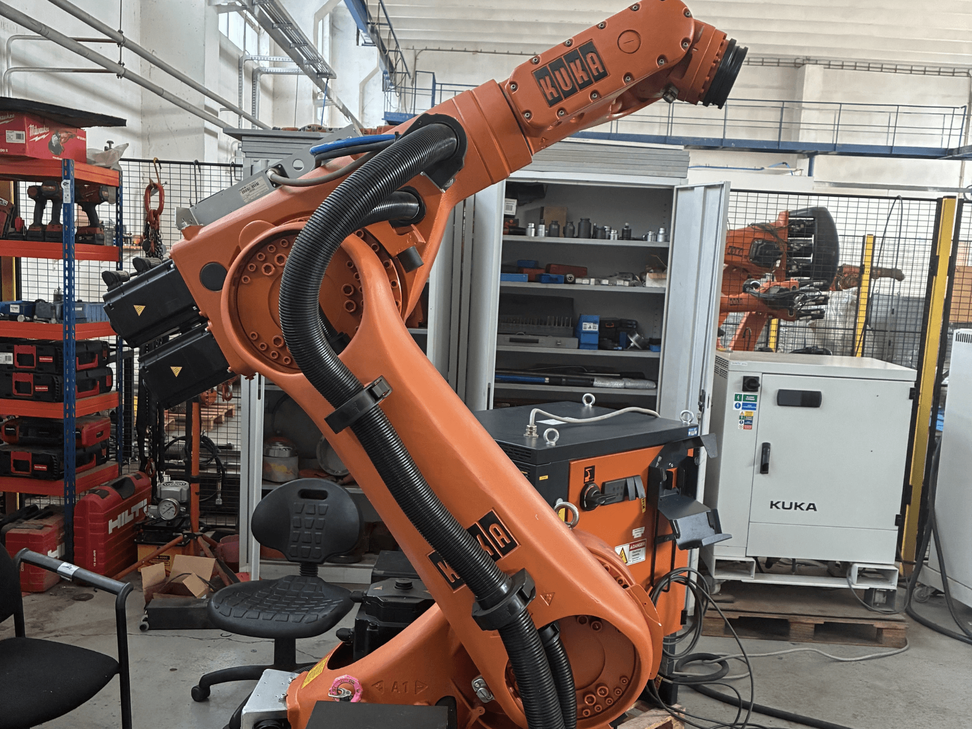KUKA KR30-3 roboto ranka pramoninėje aplinkoje, šoninis vaizdas, kuriame matomi kabeliai ir valdikliai, fone - įrankiai ir lentynos.