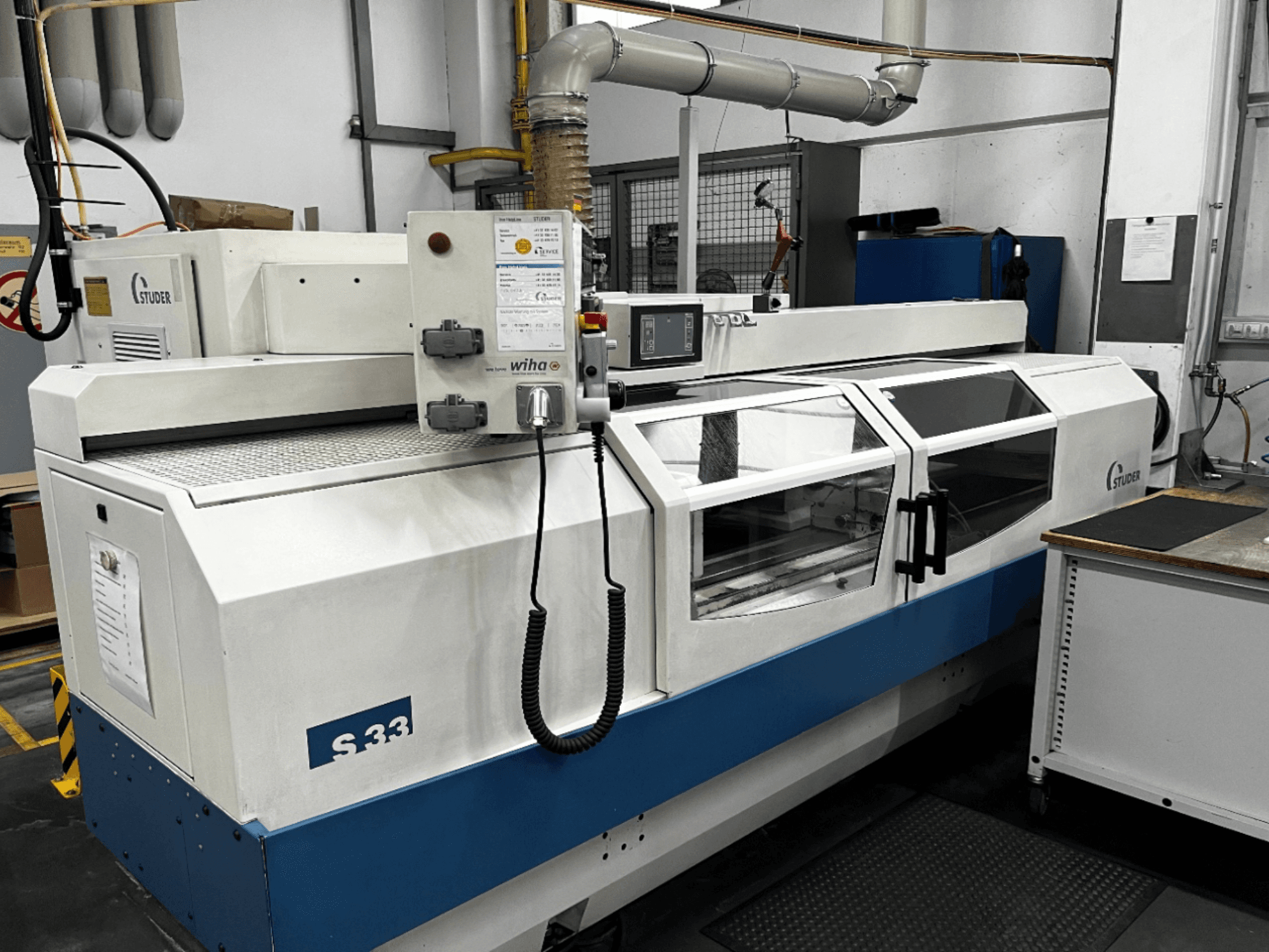 STUDER S33 CNC cilindrinio šlifavimo staklės, vaizdas iš priekio; valdymo skydelis ir stiklinės apsauginės durys. Pramoninė aplinka.