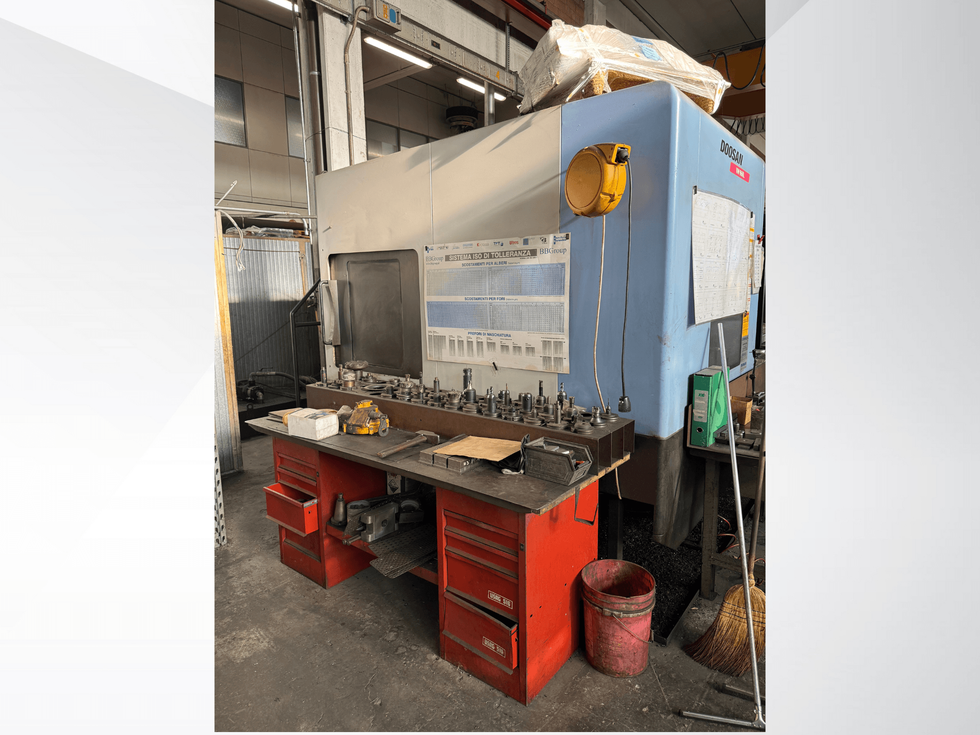 "Doosan" apdirbimo centras su raudonu darbo stalu ir saugykla, kurioje rodomi įrankiai ir įranga, žiūrint iš priekio.