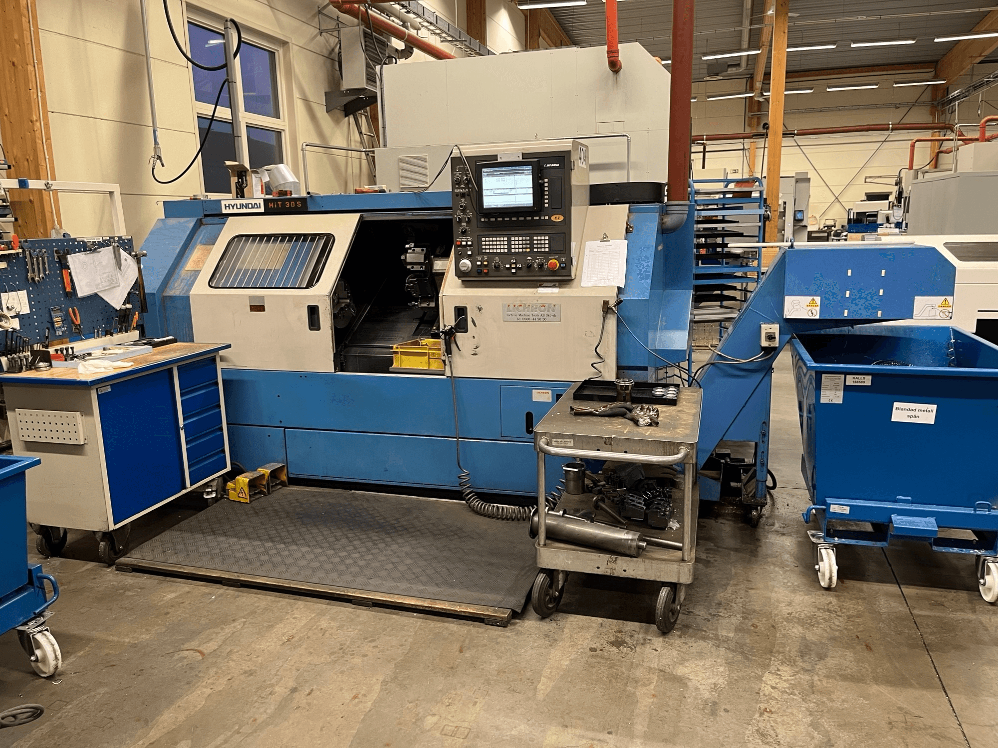 Horizontaliojo tekinimo staklės "Hyundai HIT 30S", vaizdas iš priekio, su valdymo skydeliu ir įrankiais ant šalia esančio vežimėlio dirbtuvėse.