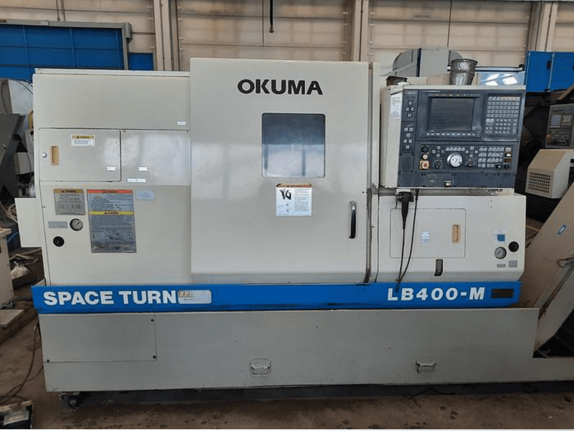 Okuma LB400-M CNC tekinimo staklės, priekinis vaizdas su valdymo skydeliu, įspėjamosiomis etiketėmis ir matomu mėlynu "SPACE TURN" prekės ženklu.
