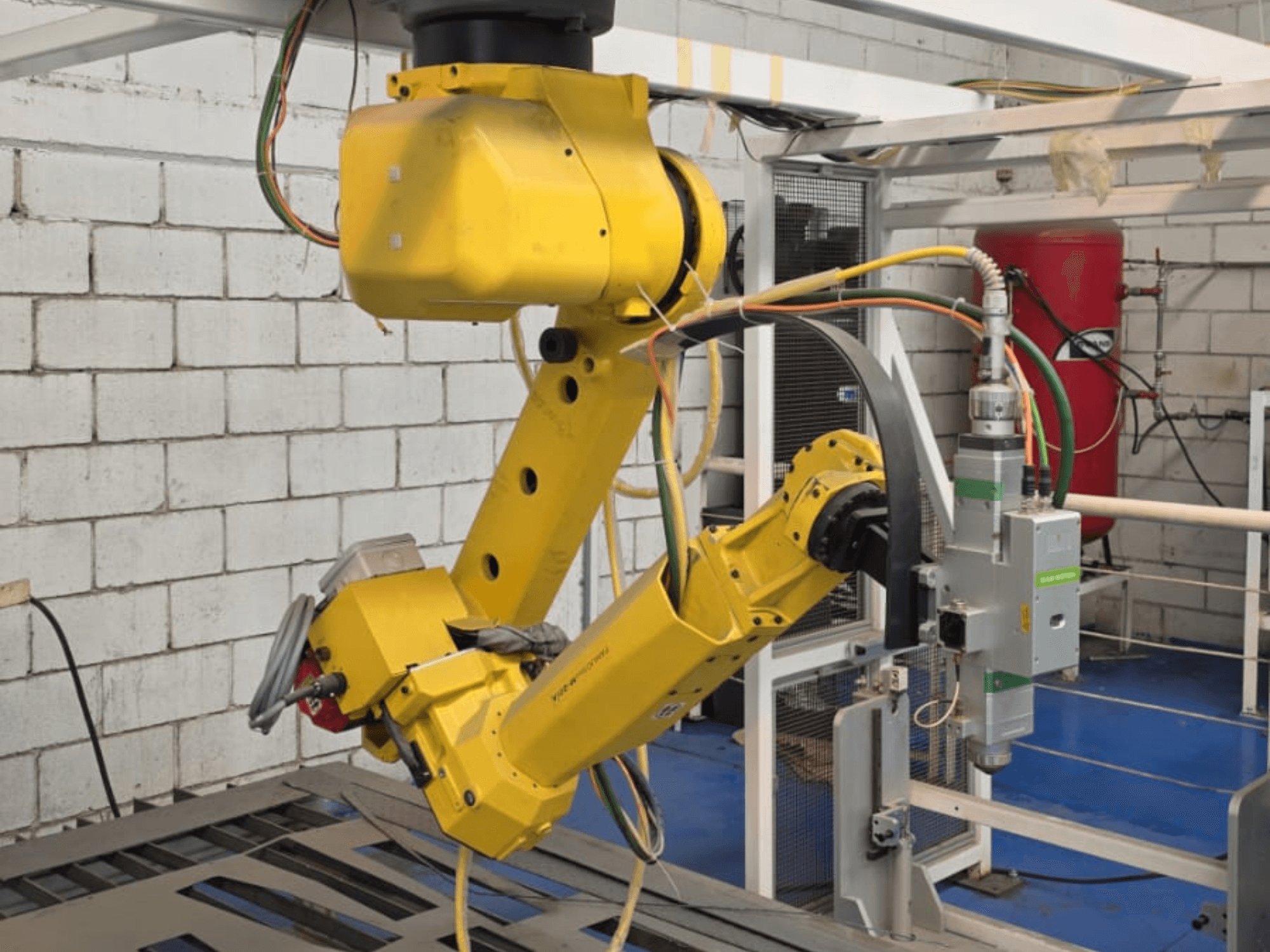 Geltonos spalvos roboto ranka su tiksliu įrankiu, turinti daugybę sąnarių ir kabelių, žiūrint iš priekio pramoninėje aplinkoje.