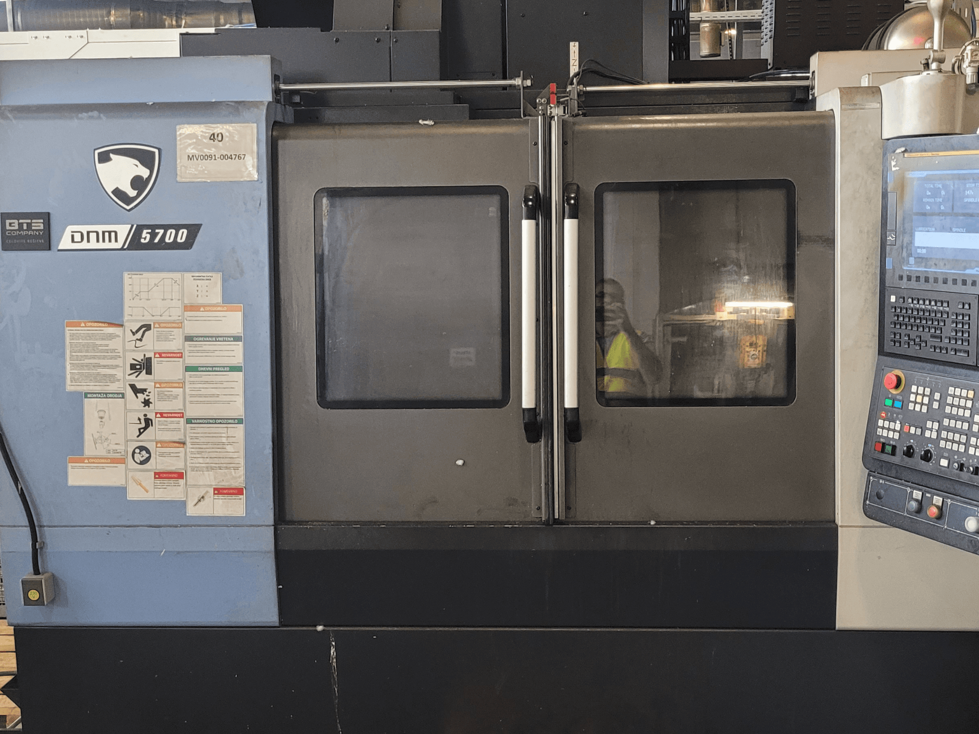 „DOOSAN DNM 5700“ vertikalaus apdirbimo centro vaizdas iš priekio su valdymo pultu, saugos etiketėmis ir šiek tiek praviromis durimis.