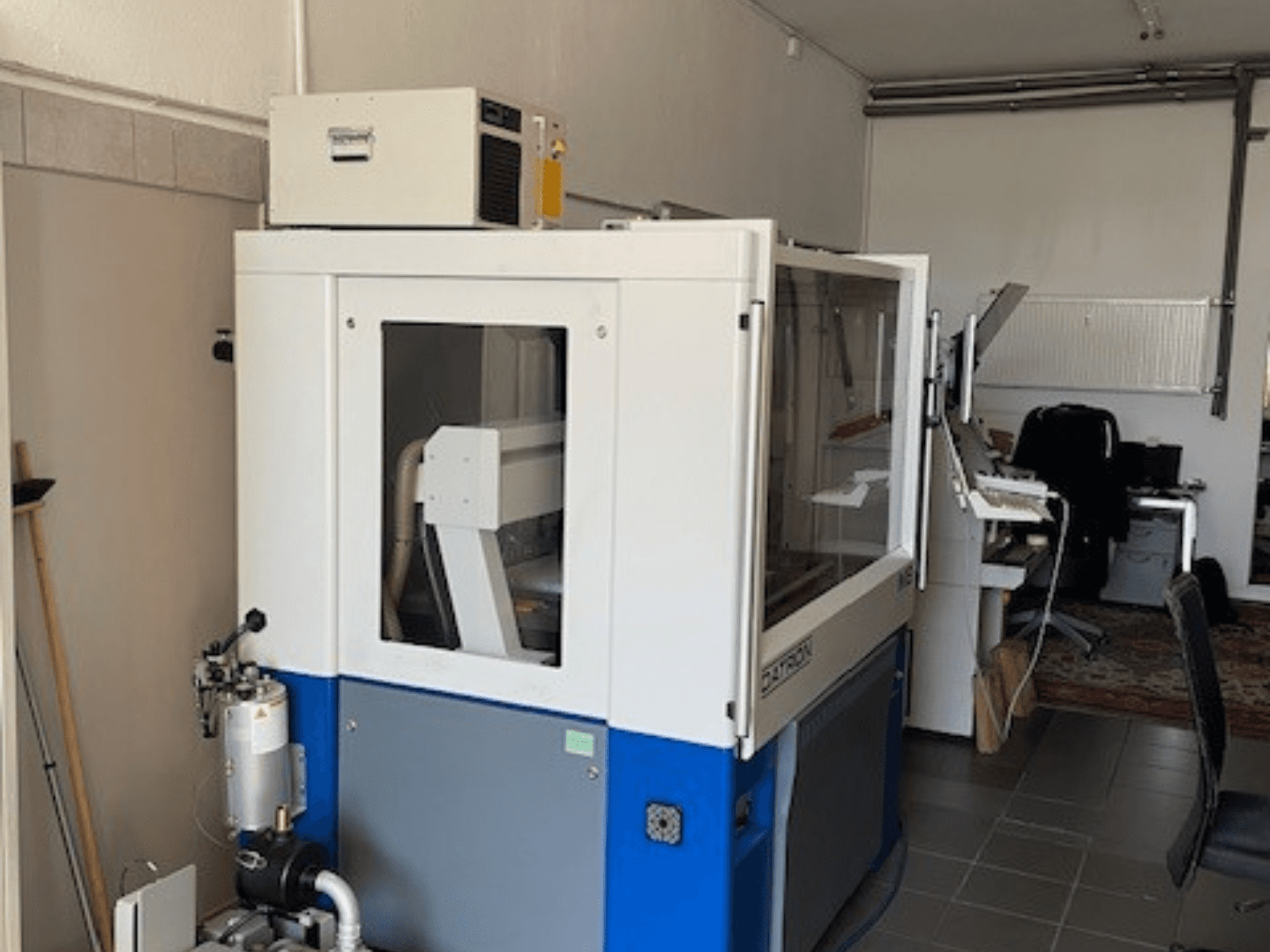 Mėlynos ir baltos spalvos pramoninė mašina su permatomais skydeliais, vaizdas iš priekio, yra valdymo prieiga ir matomas vėdinimo įrenginys.