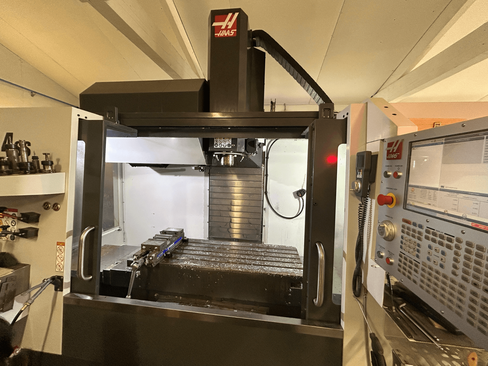 HAAS VF-4SS vertikalusis apdirbimo centras, vaizdas iš priekio, matomas valdymo skydelis, įrankių laikiklis ir darbo zona su metalo drožlėmis.