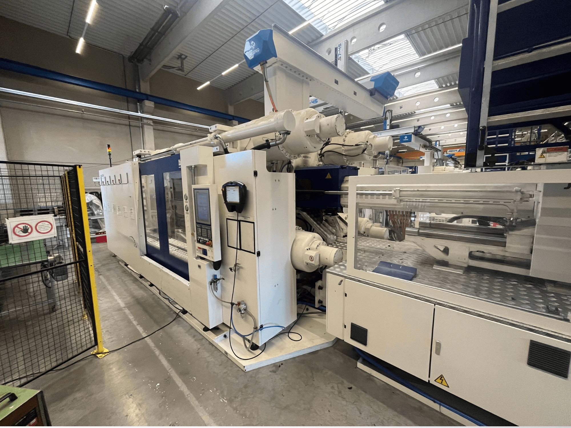 Baltos ir mėlynos spalvos liejimo mašina, vaizdas iš priekio, su valdymo pultu, roboto ranka ir saugos ženklais.