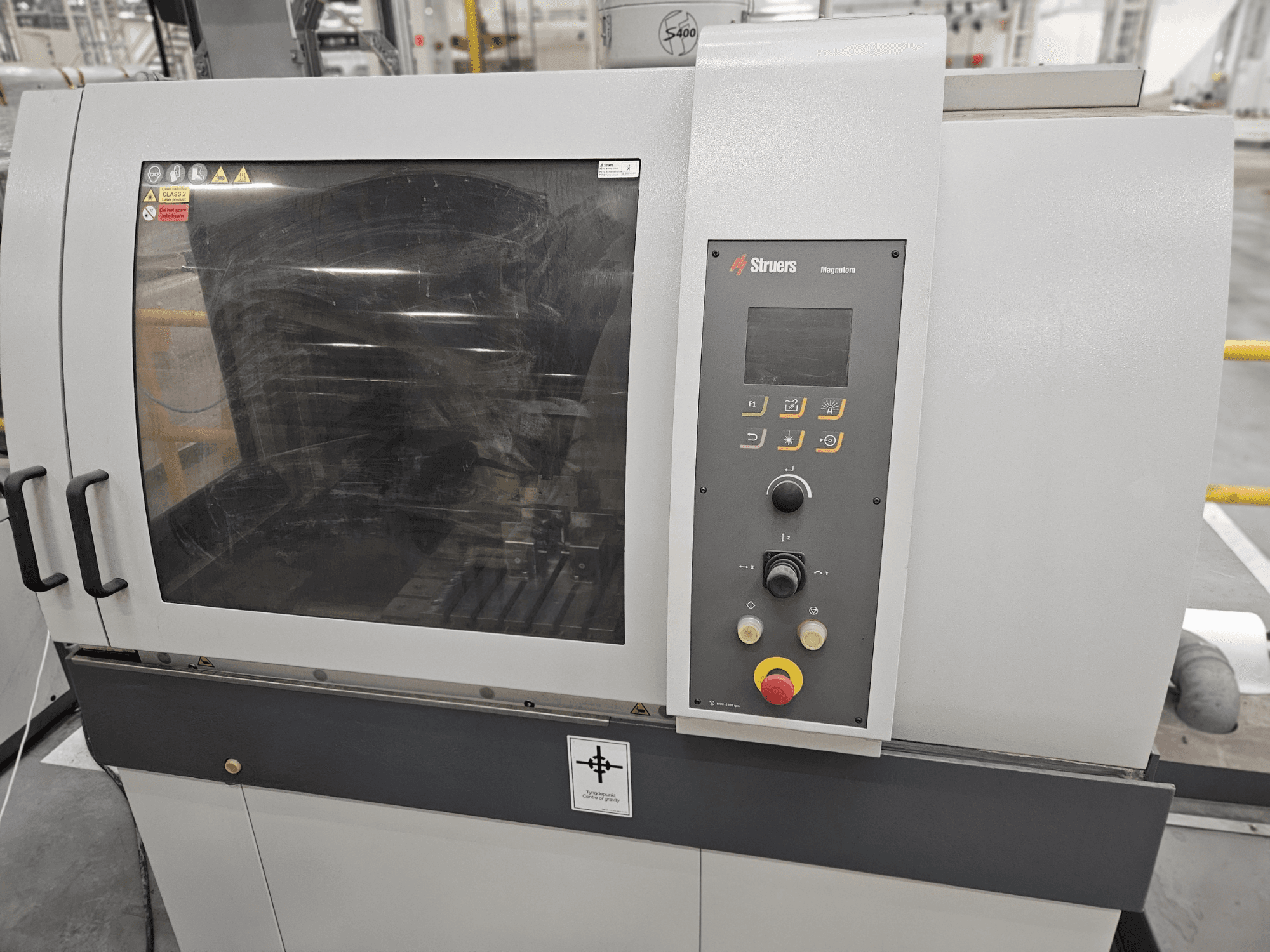 "Struers Magpentor" mašinos priekinis vaizdas su valdymo skydeliu, mygtukais ir dideliu stebėjimo langu. Matomos techninės etiketės.
