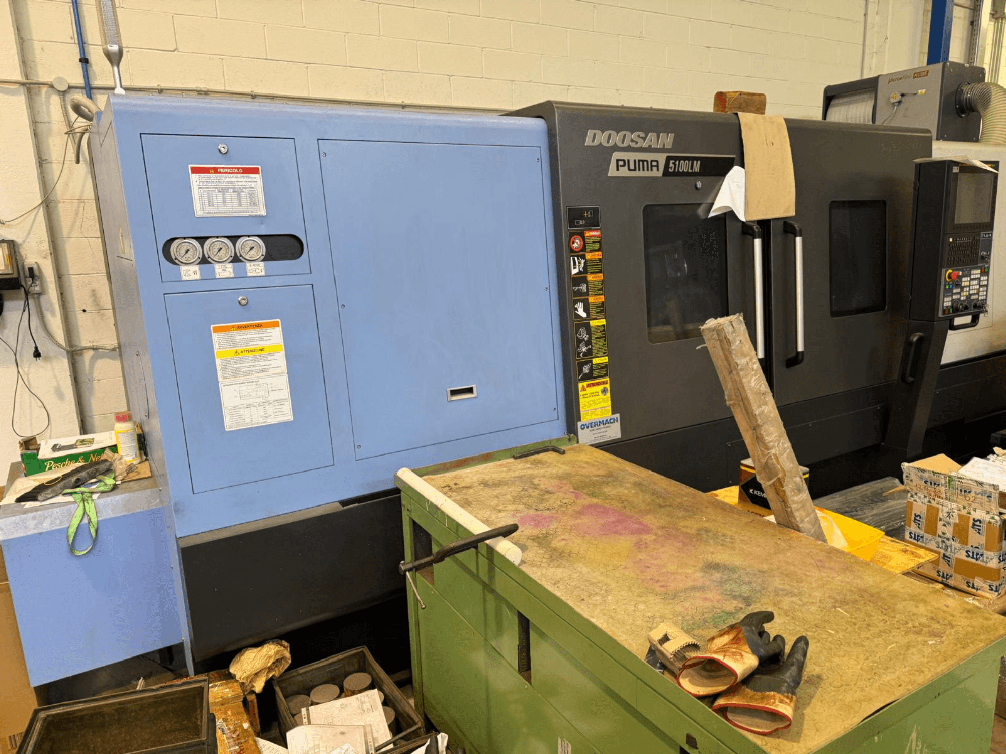 CNC staklės "Doosan Puma 5100LM" ir valdymo pultas, šoninis vaizdas. Ant mėlynojo valdymo bloko matomi matuokliai ir įspėjamosios etiketės.