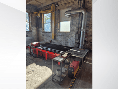 Raudonas CNC plazminio pjovimo stalas su valdymo pultu ir metaliniu išmetimo kanalu, žiūrint iš šono gerai apšviestose dirbtuvėse.