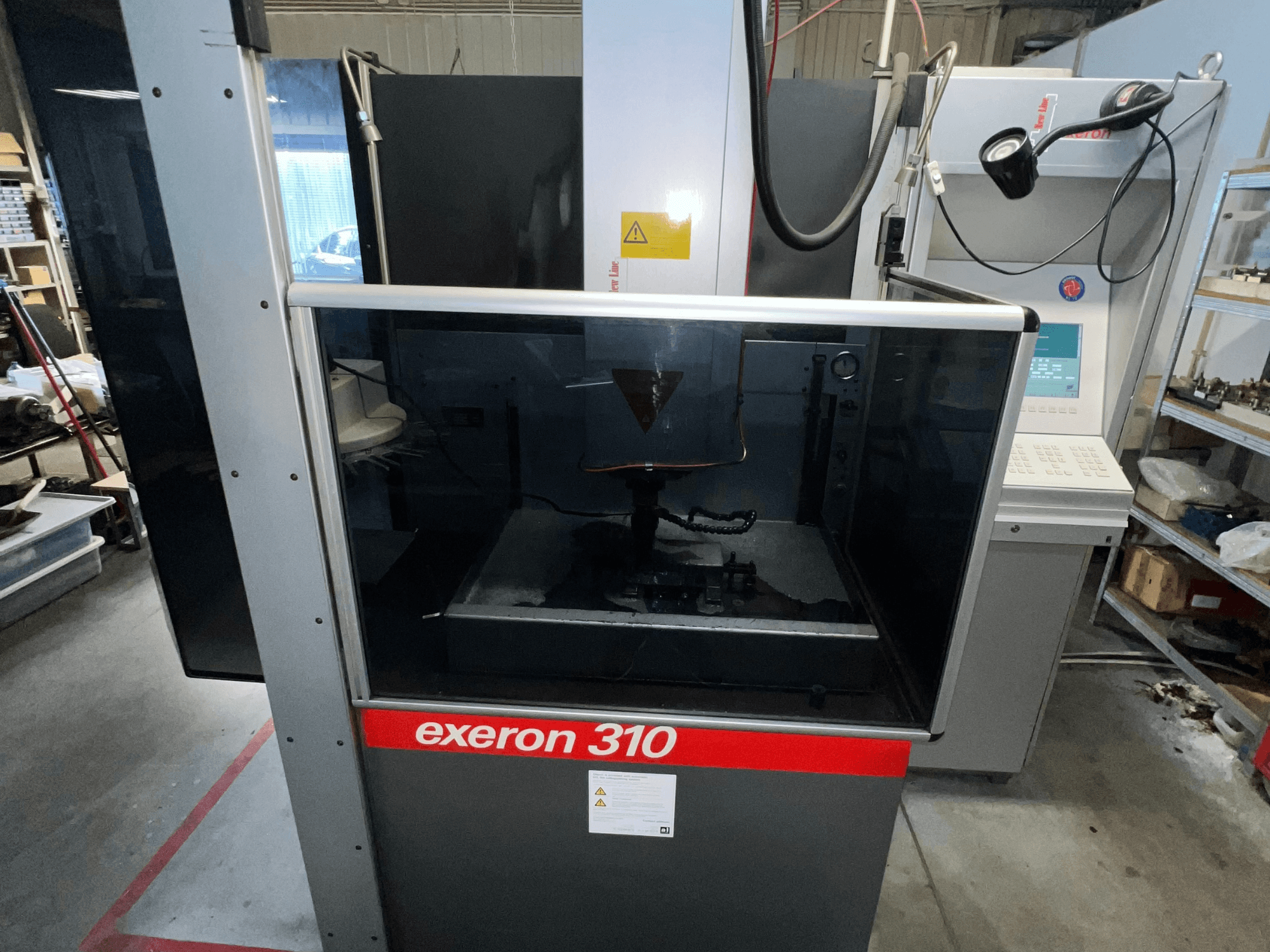 "Exeron 310" mašinos vaizdas iš priekio: stiklinė kamera ir valdymo skydelis bei darbo vietos lentynos.