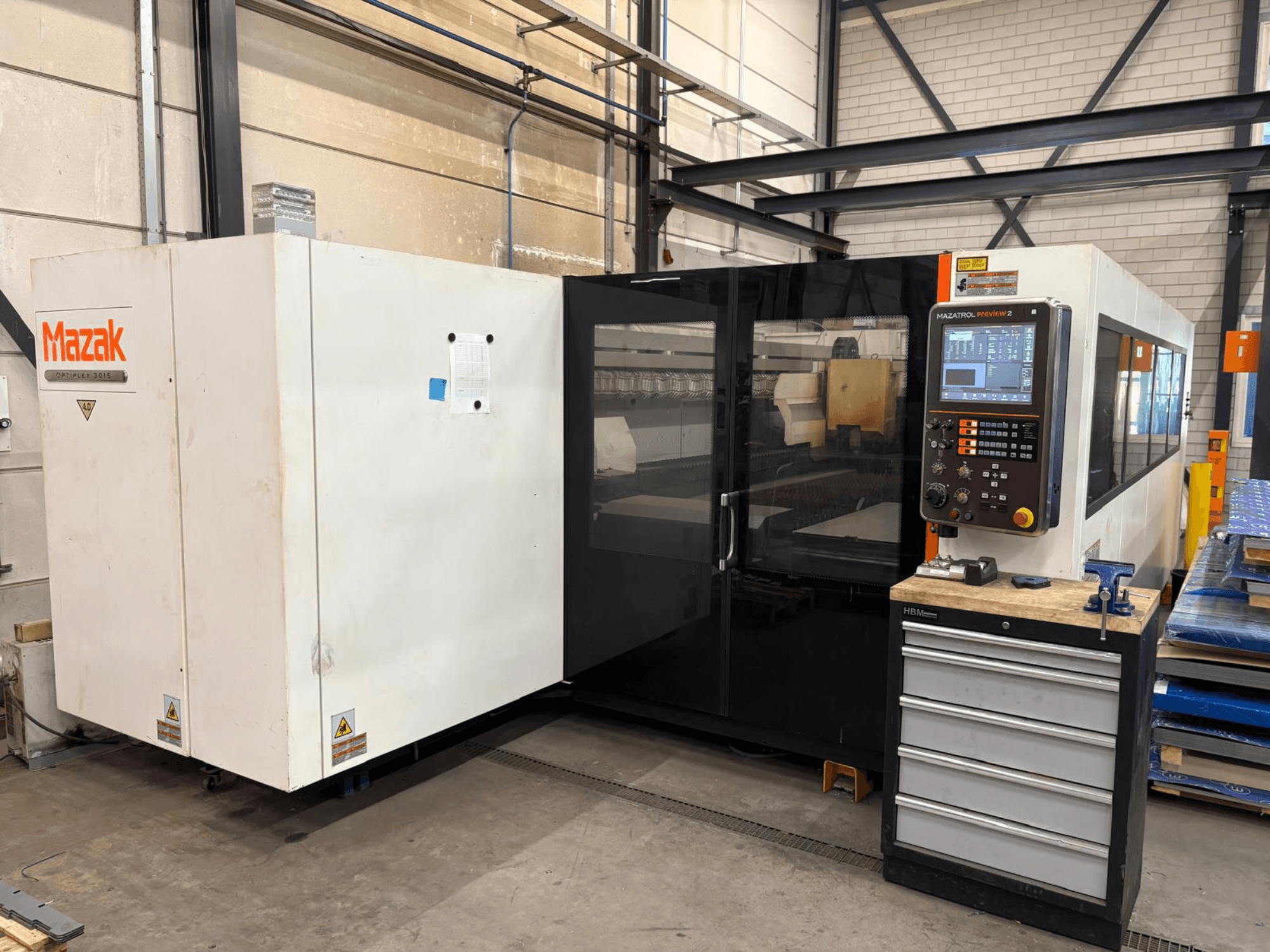 „Mazak Optiplex 3015“ CO₂ lazerinio pjovimo staklės, vaizdas iš priekio, kuriame matomas valdymo pultas ir pramoninė aplinka.