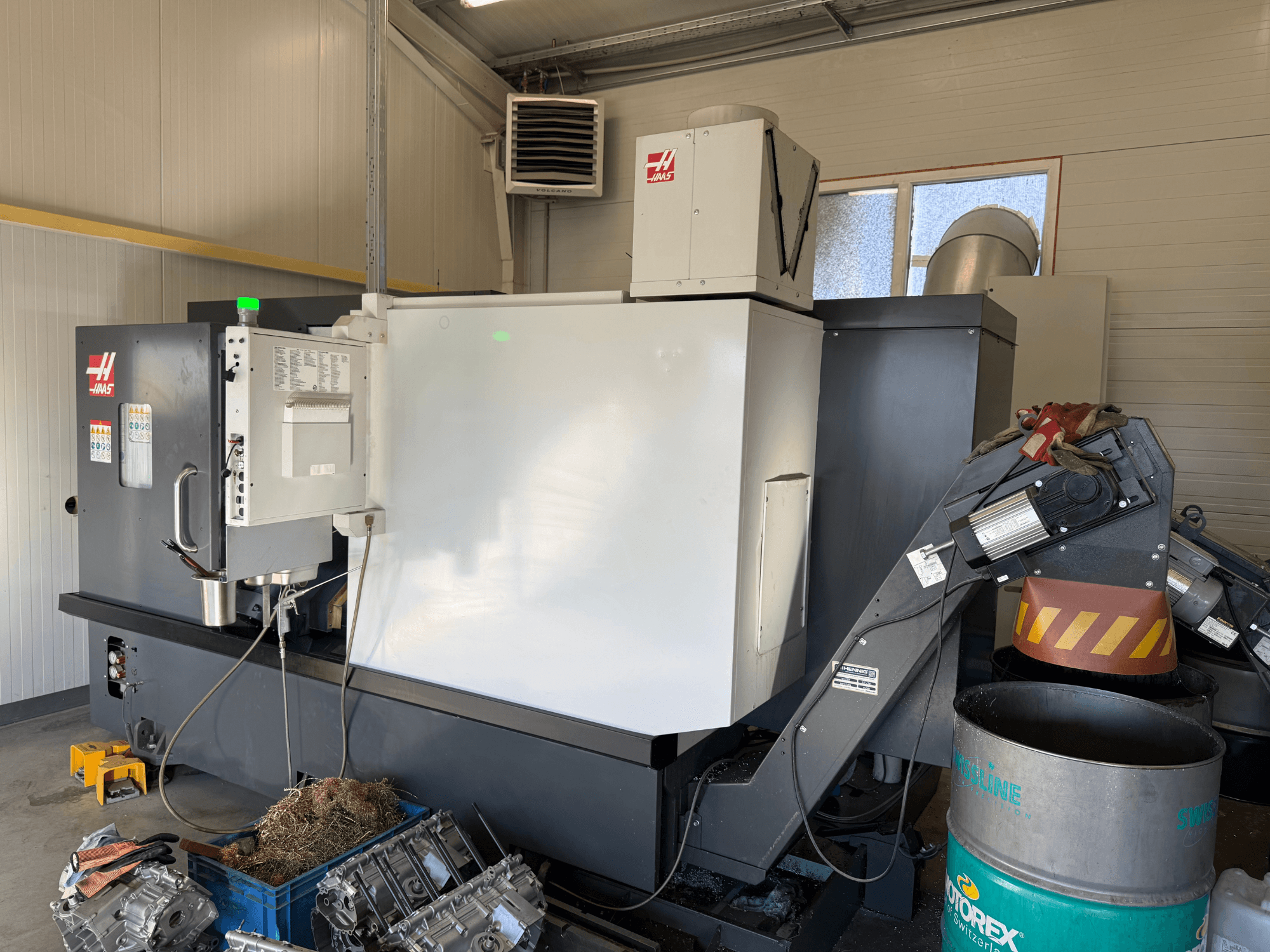 "Haas" CNC staklės su valdymo skydeliu, vaizdas iš šono; matomos aušinimo skysčio ir alyvos linijos; metalo atliekos mėlynoje šiukšliadėžėje; pramoninė aplinka.