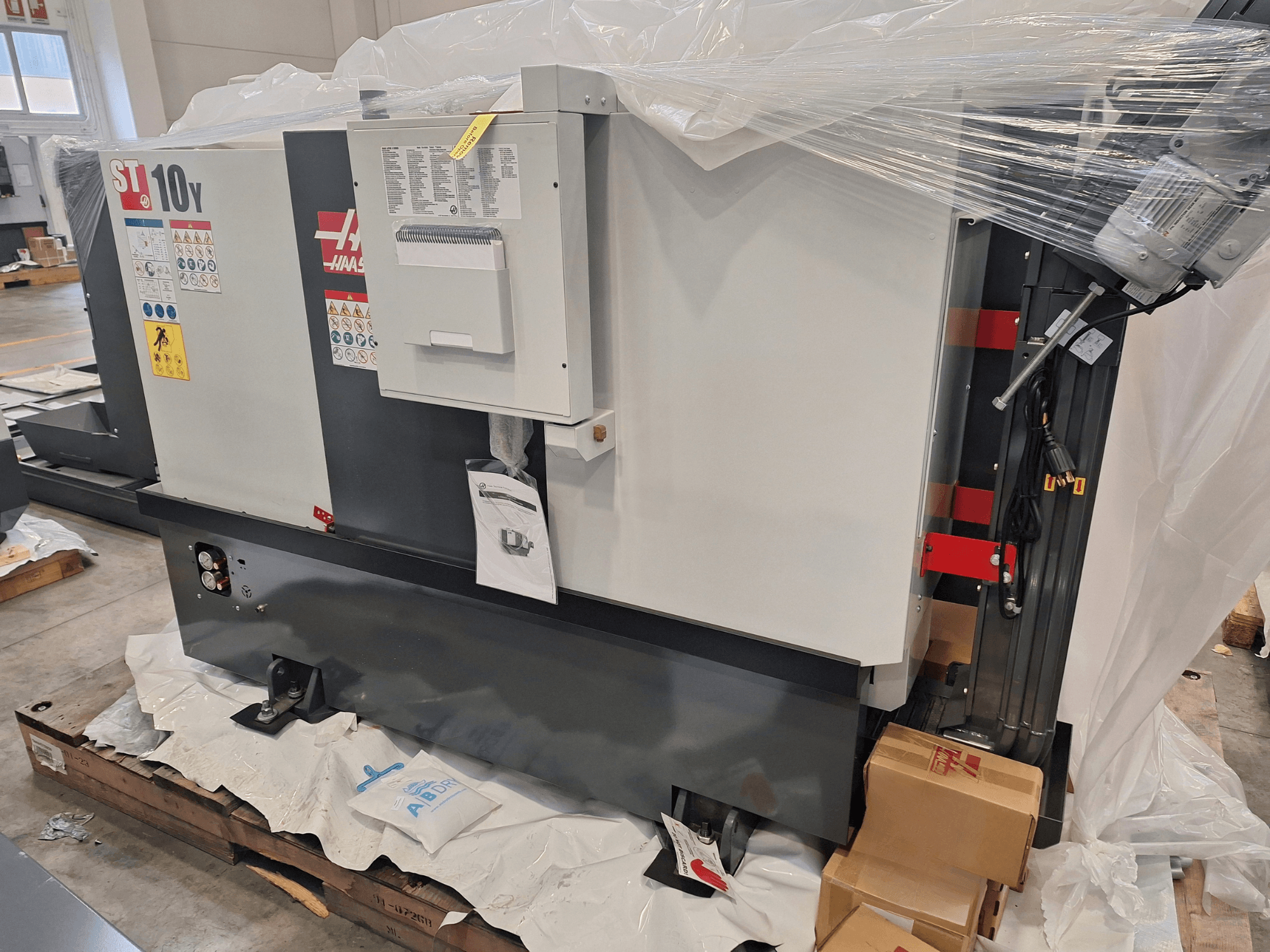 HAAS ST-10Y EU horizontalaus tekinimo staklių vaizdas iš šono, įvyniojamos į plastikinę plėvelę, matomas valdymo pultas ir įspėjamieji užrašai.