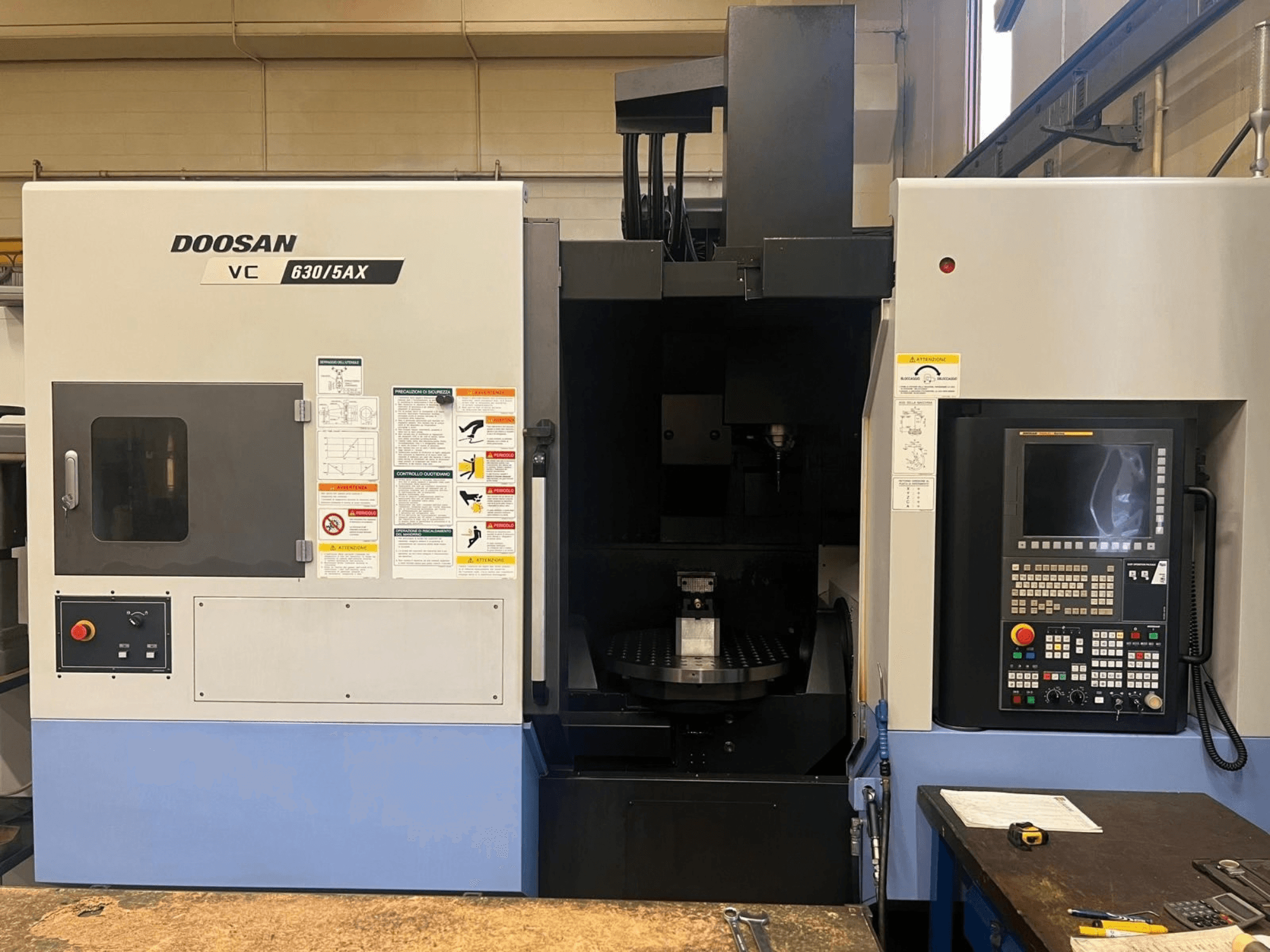 „DOOSAN VC 630/5AX“ vertikaliojo apdirbimo centro vaizdas iš priekio, kuriame matomas valdymo pultas ir suklio sritis su aiškiai matomomis saugos etiketėmis.