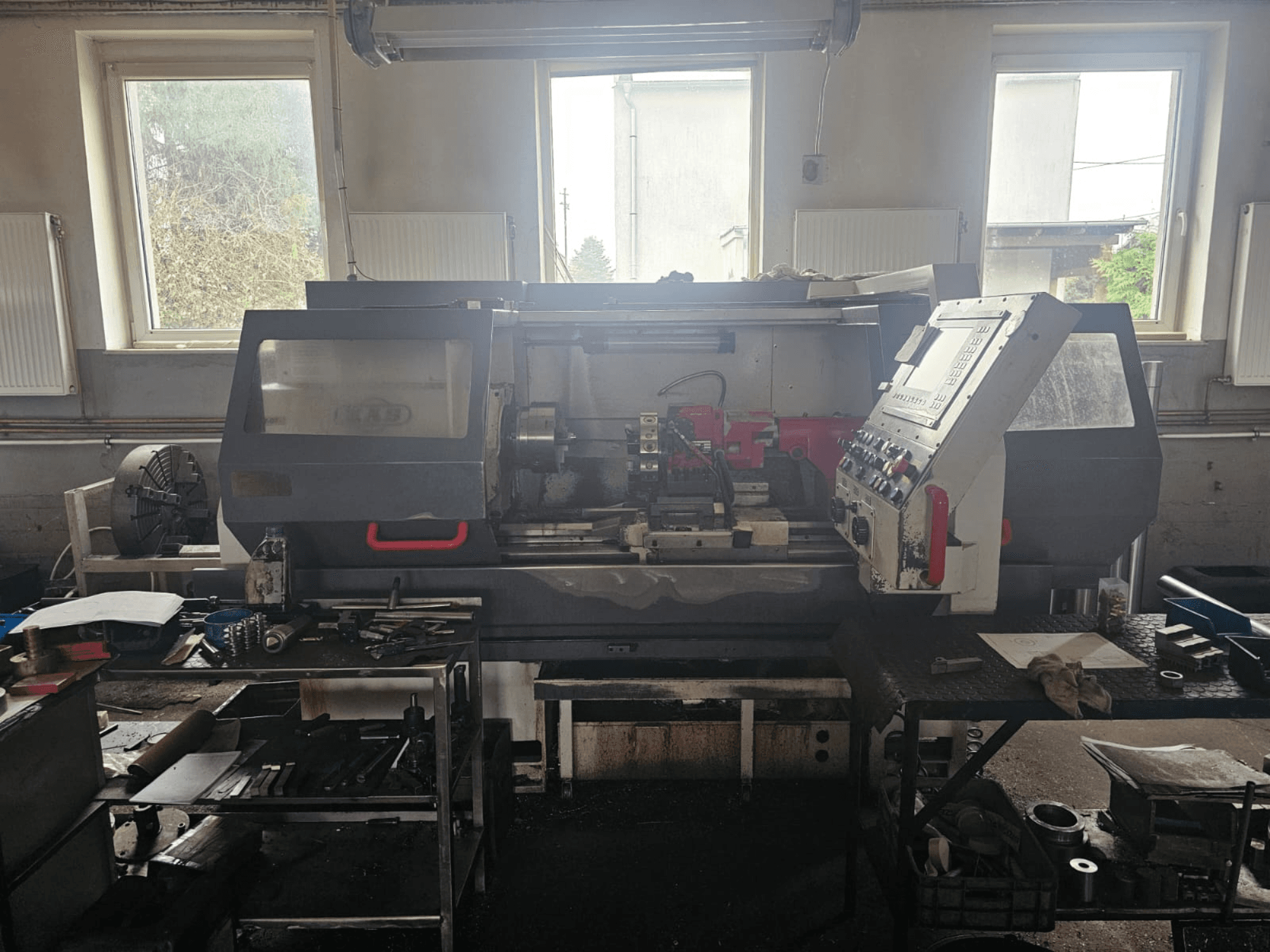 Tekinimo staklės dirbtuvėse, vaizdas iš priekio, su valdymo pultu ir įrankiais ant stalų, apsuptos langų ir užgriozdintos darbo vietos.