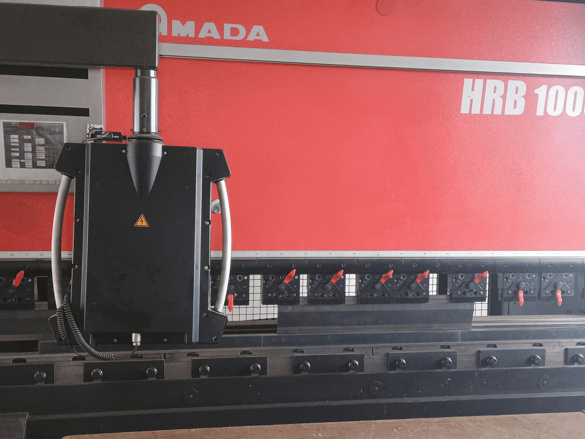 "Amada HRB 100" lenkimo staklių vaizdas iš priekio; juoda valdymo galvutė su kūginiu antgaliu ir reguliuojamais spaustuvais.