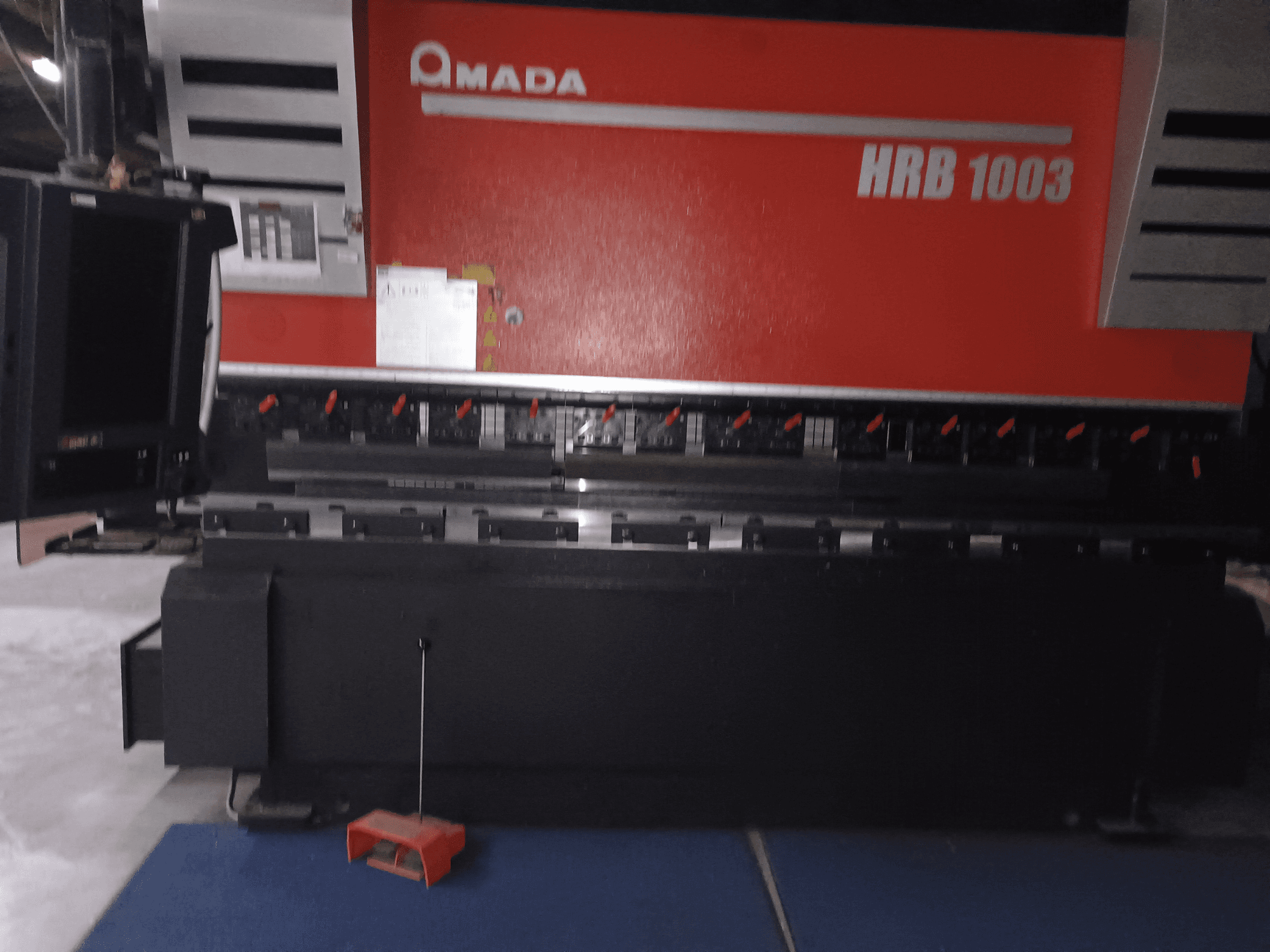 "Amada HRB 1003" presinis stabdys, vaizdas iš priekio; valdymo skydelis su raudonomis rankenėlėmis ir juodu korpusu, įrengtas pramoninėje aplinkoje.