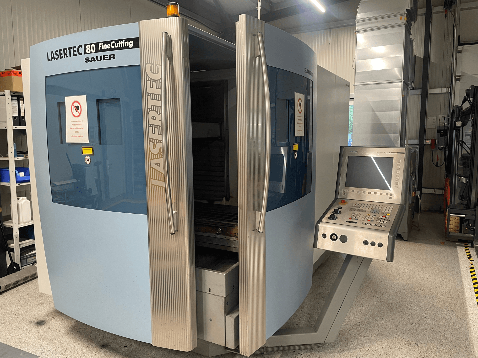 DMG Sauer „Lasertec80 FineCutting“ lakštinio metalo apdirbimo staklių vaizdas iš priekio su valdymo pultu, atidarytomis durimis ir matomais saugos ženklais.