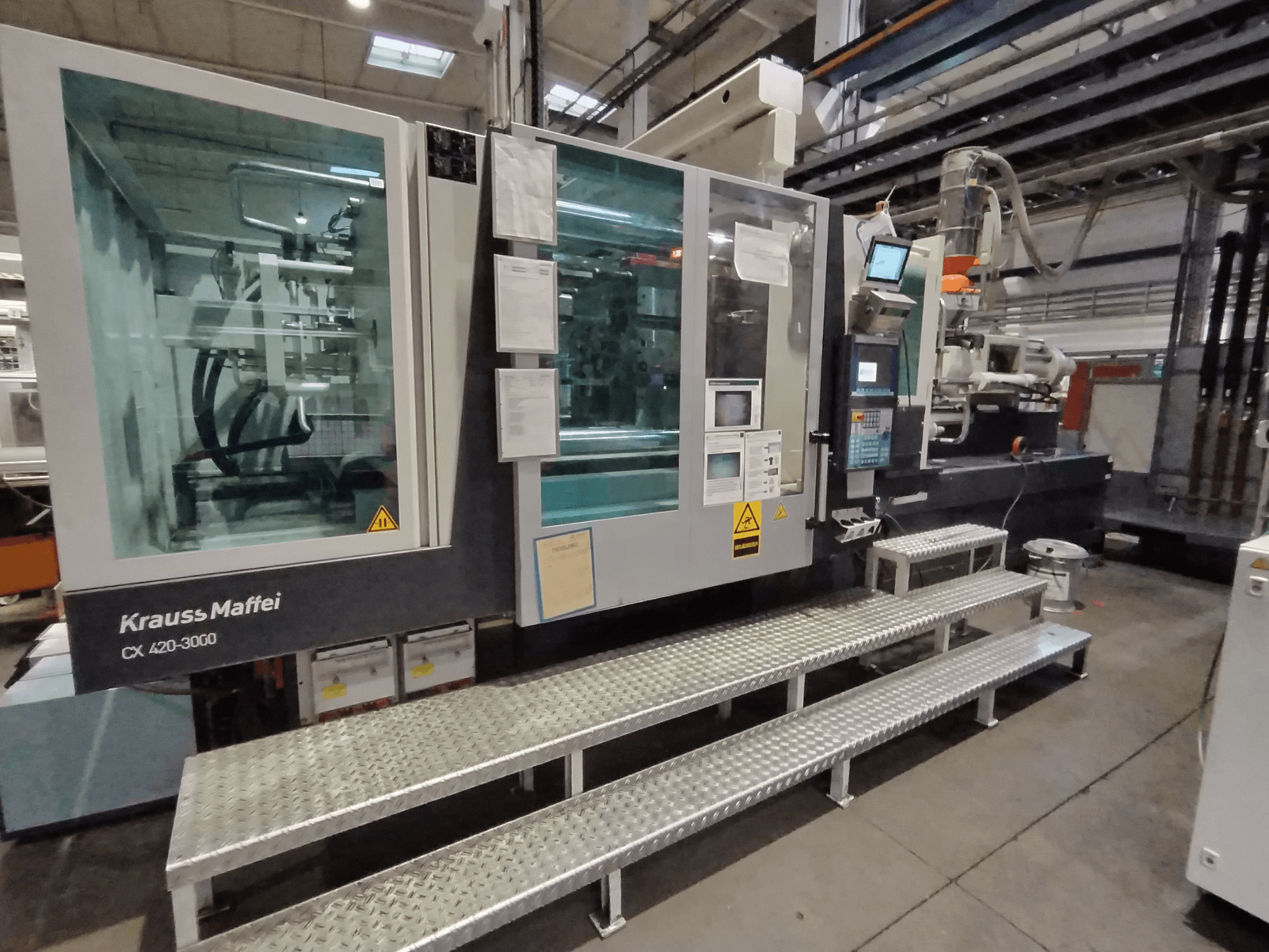 "Krauss Maffei CX 420-3000" šoninis vaizdas, matomas valdymo skydelis ir saugos ženklai, pastatytas ant metalinių laiptelių.