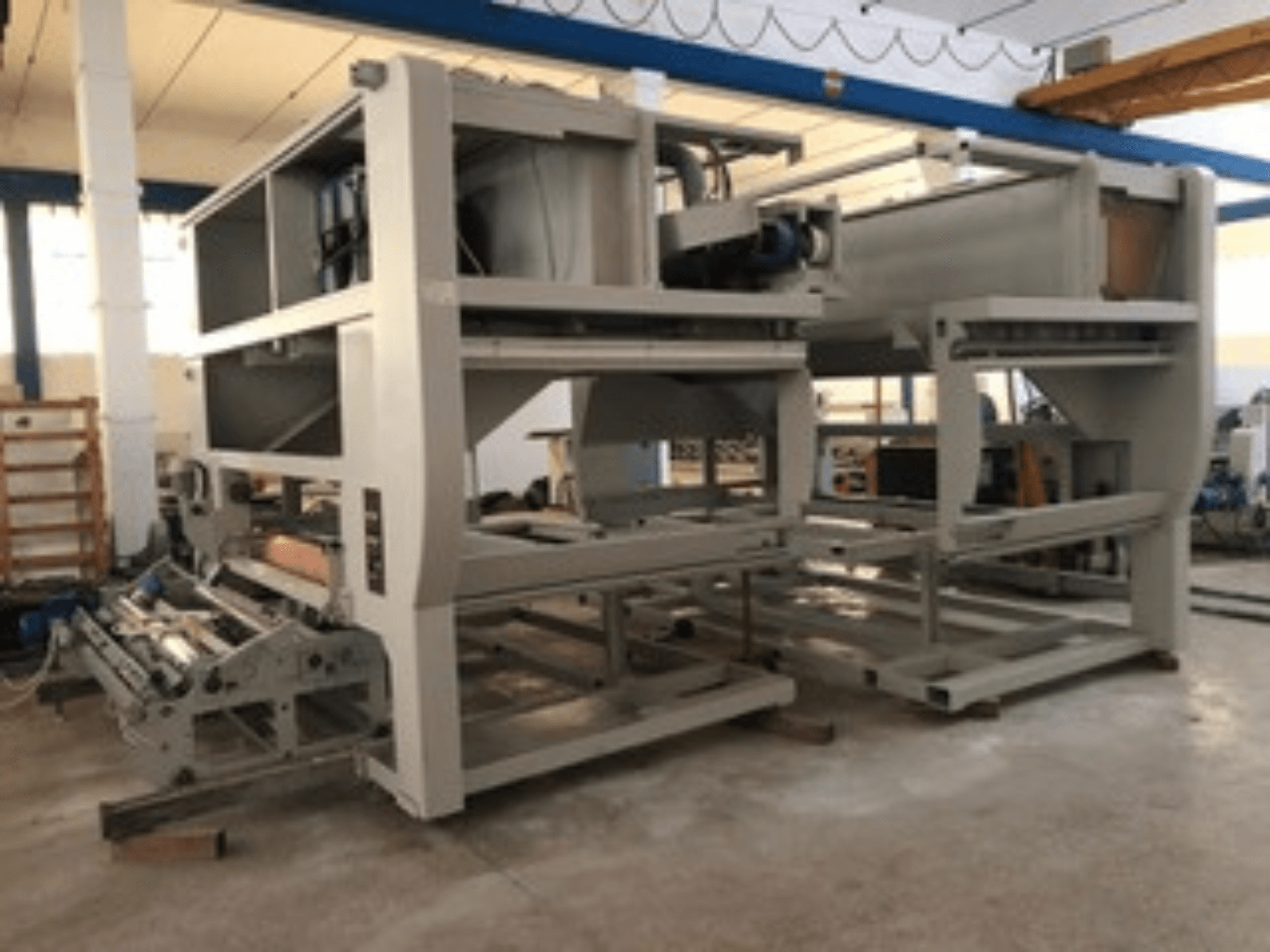 Pramoninė mašina su keliais konvejerio ritinėliais ir didele konstrukcija; vaizdas iš šono, pilka spalva, gerai apšviestoje gamykloje.