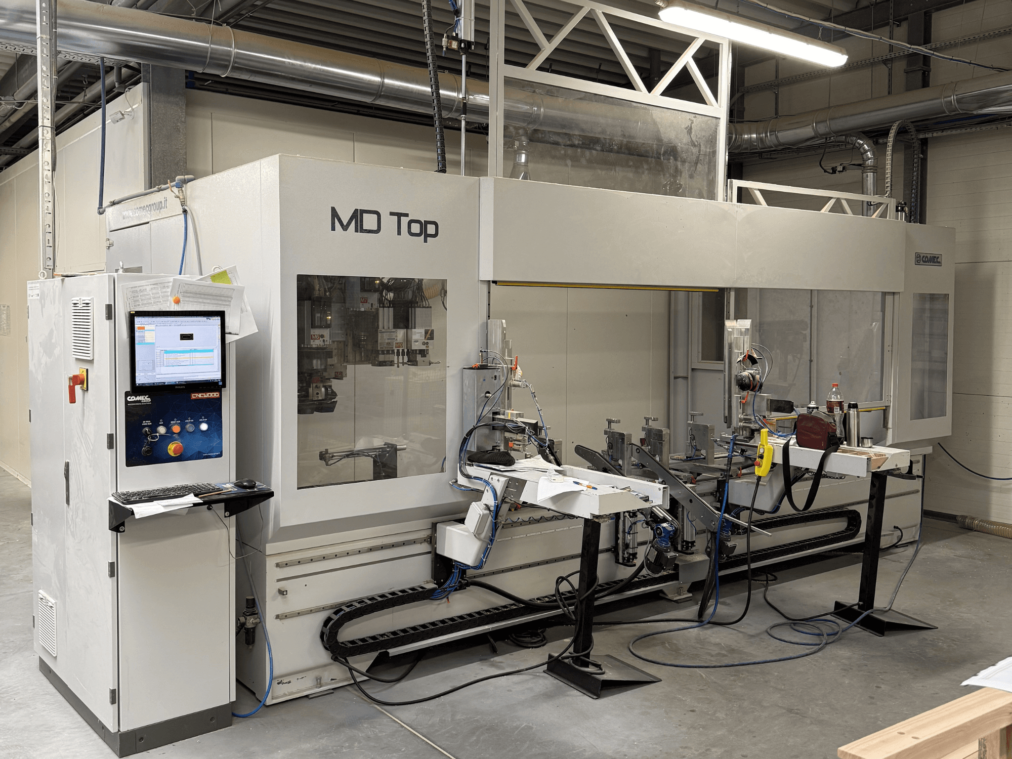 MD Top CNC staklės, žiūrint iš priekio, su valdymo pultu, roboto rankomis ir darbo vieta su įrankiais bei medžiagomis.