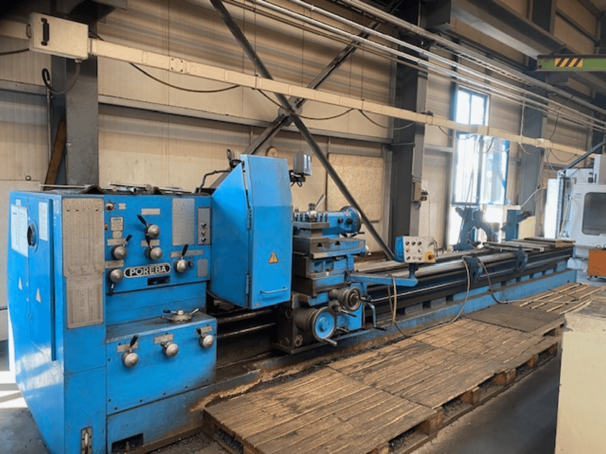 Mėlynos spalvos POREBA tekinimo staklės su valdymo rankenėlėmis, įrankių atrama ir mediniais padėklais iš priekio.