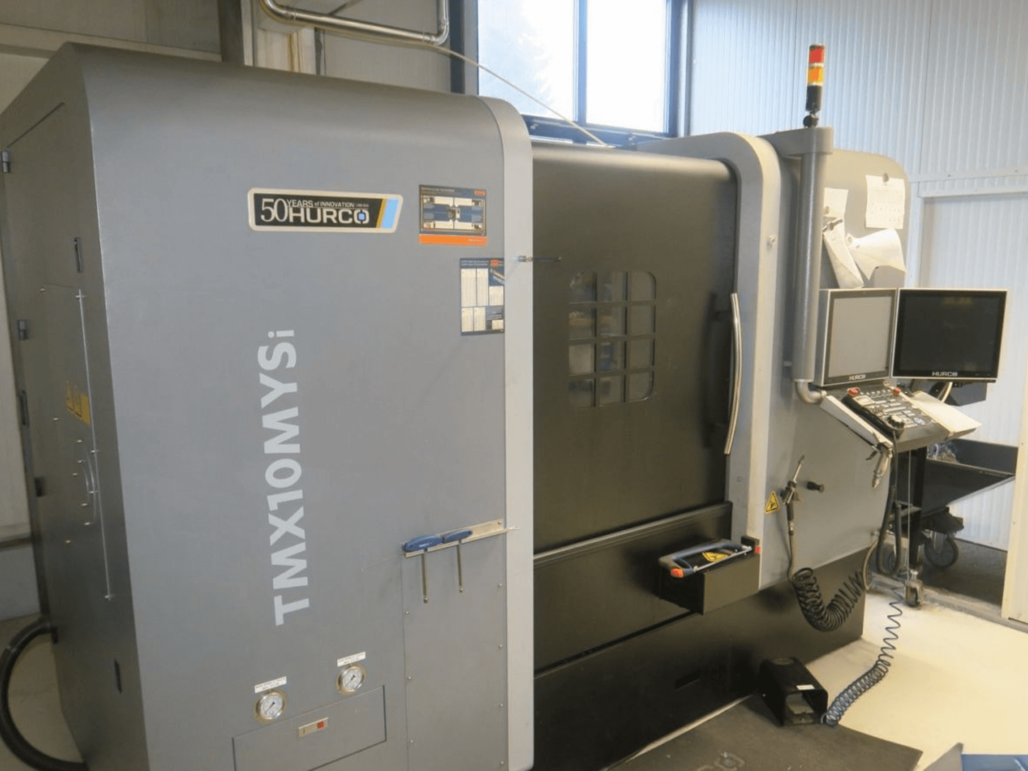 Hurco TMX10MYSi CNC staklės iš šono; rodomas valdymo skydelis, avarinės lemputės ir matuokliai.