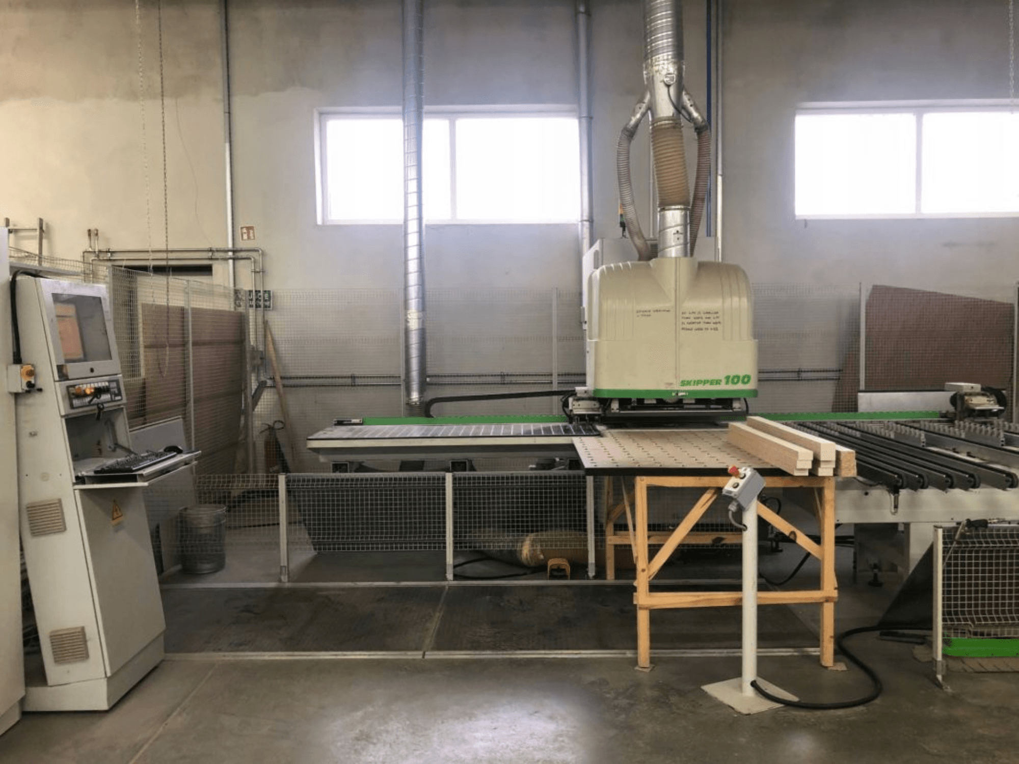 BIESSE SKIPPER 100 CNC apdirbimo centras dirbtuvėse; vaizdas iš priekio rodo stakles su valdymo pultu ir ant stalo padėtą medieną.