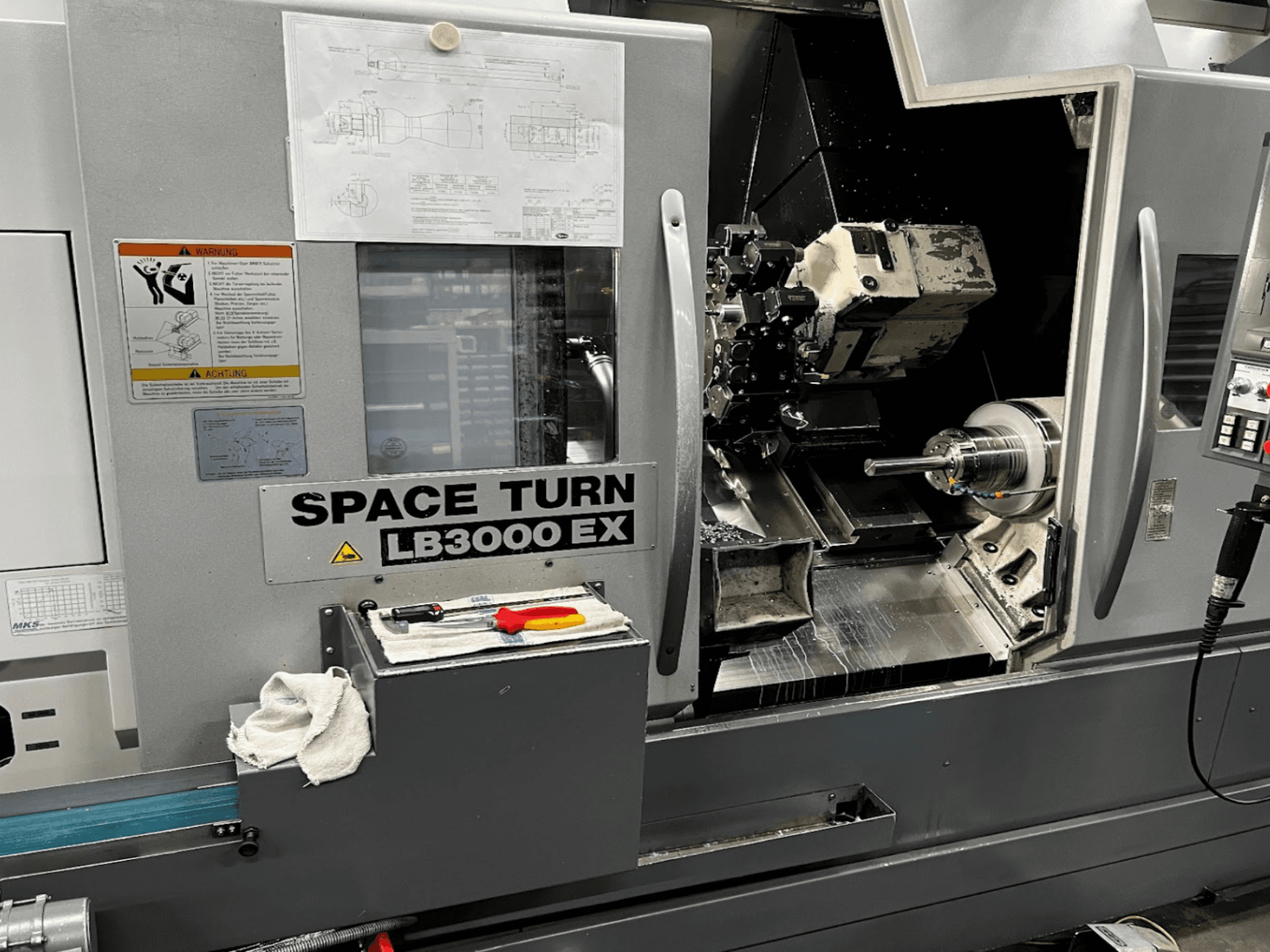 Okuma LB3000EX MYW horizontalios tekinimo staklės, priekinis vaizdas, matomas valdymo skydelis, įrankiai ir darbo vietos detalės.