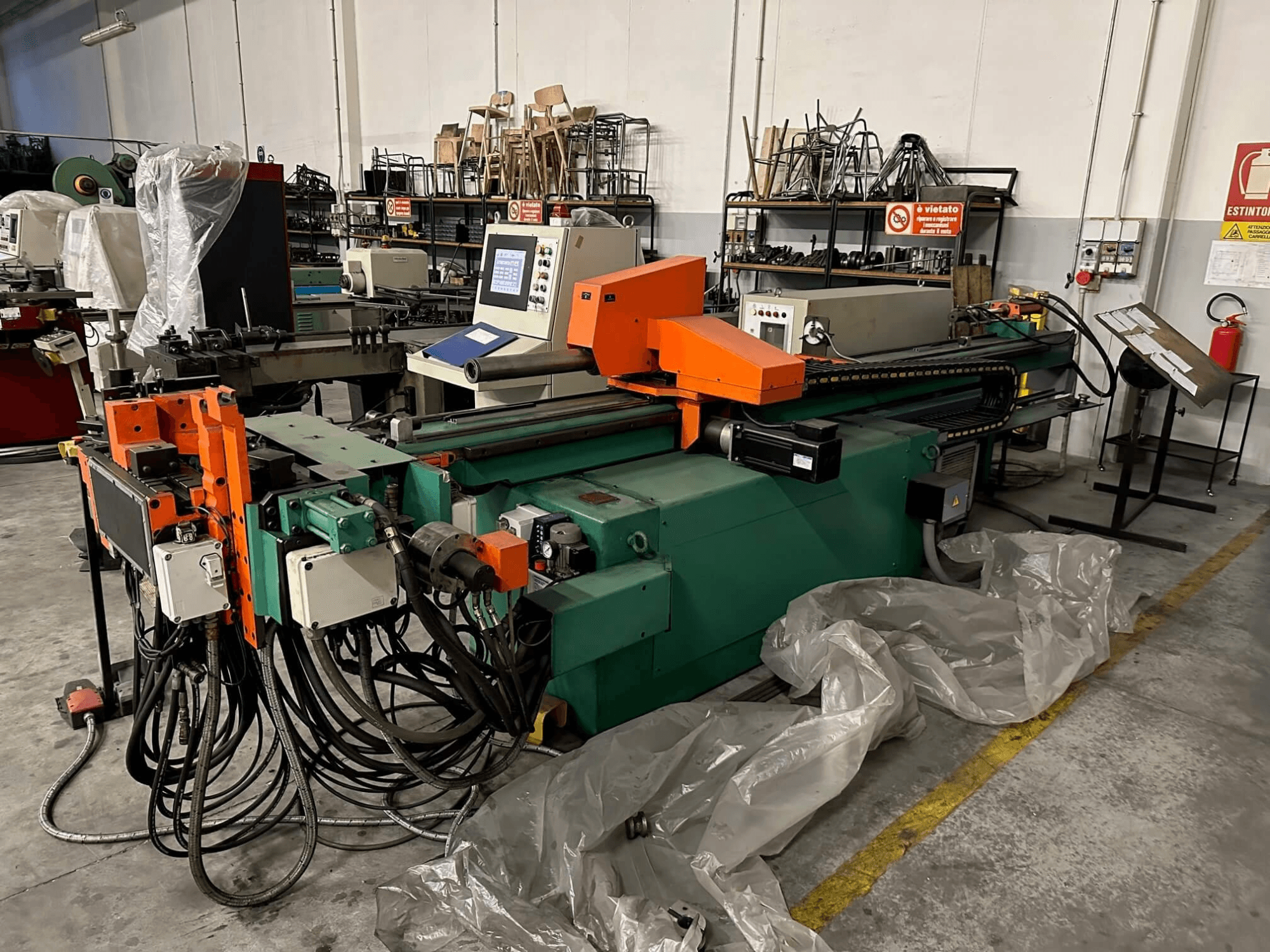 Pramoninė mašina su oranžiniu ir žaliu korpusu, valdymo skydeliu ir daugybe kabelių, žiūrint iš priekio dirbtuvėse.