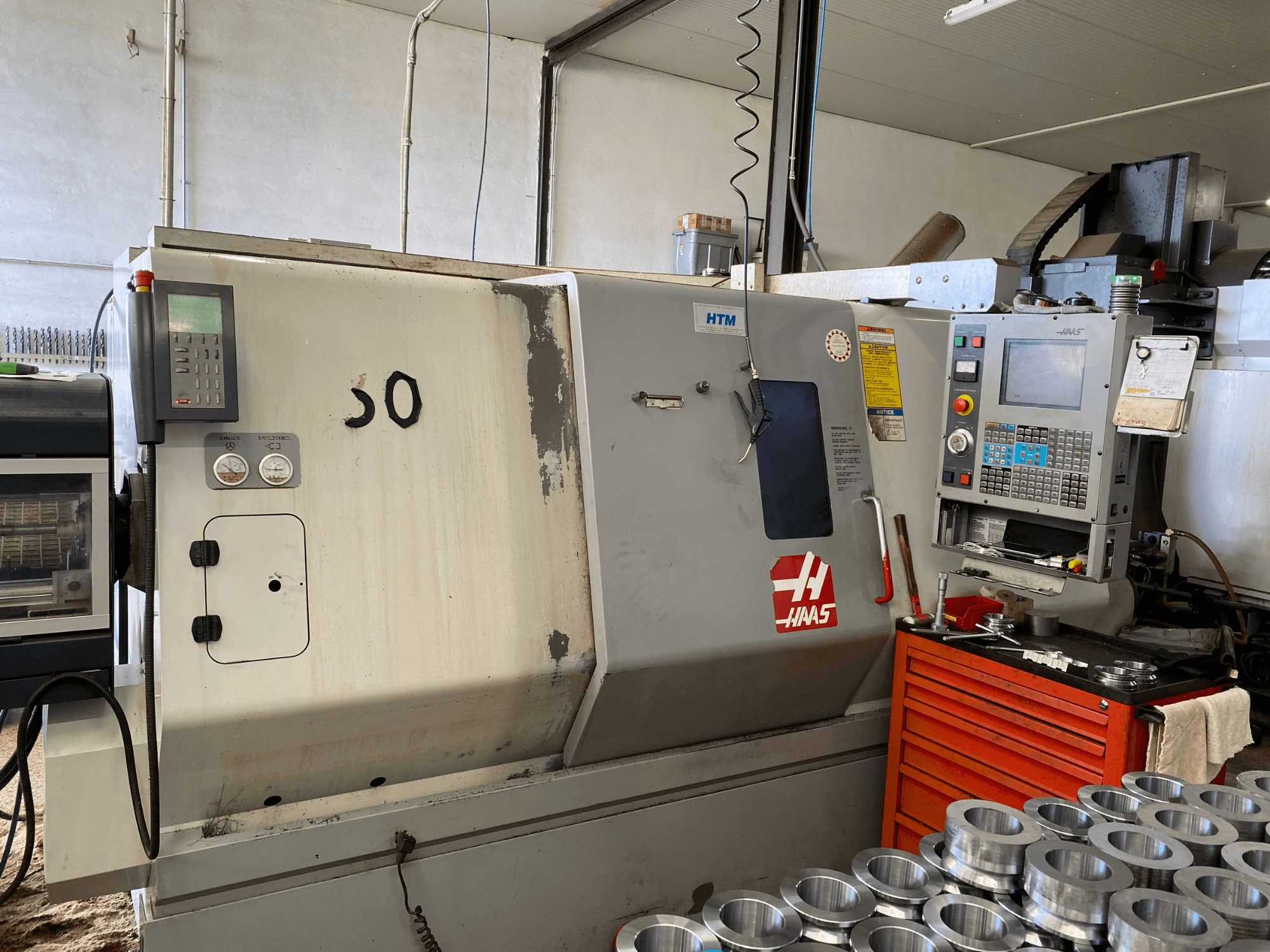 Haas CNC staklės su valdymo skydeliu, vaizdas iš priekio, kuriame matomi valdymo mygtukai, matuokliai ir šalia sukrautos metalinės detalės.