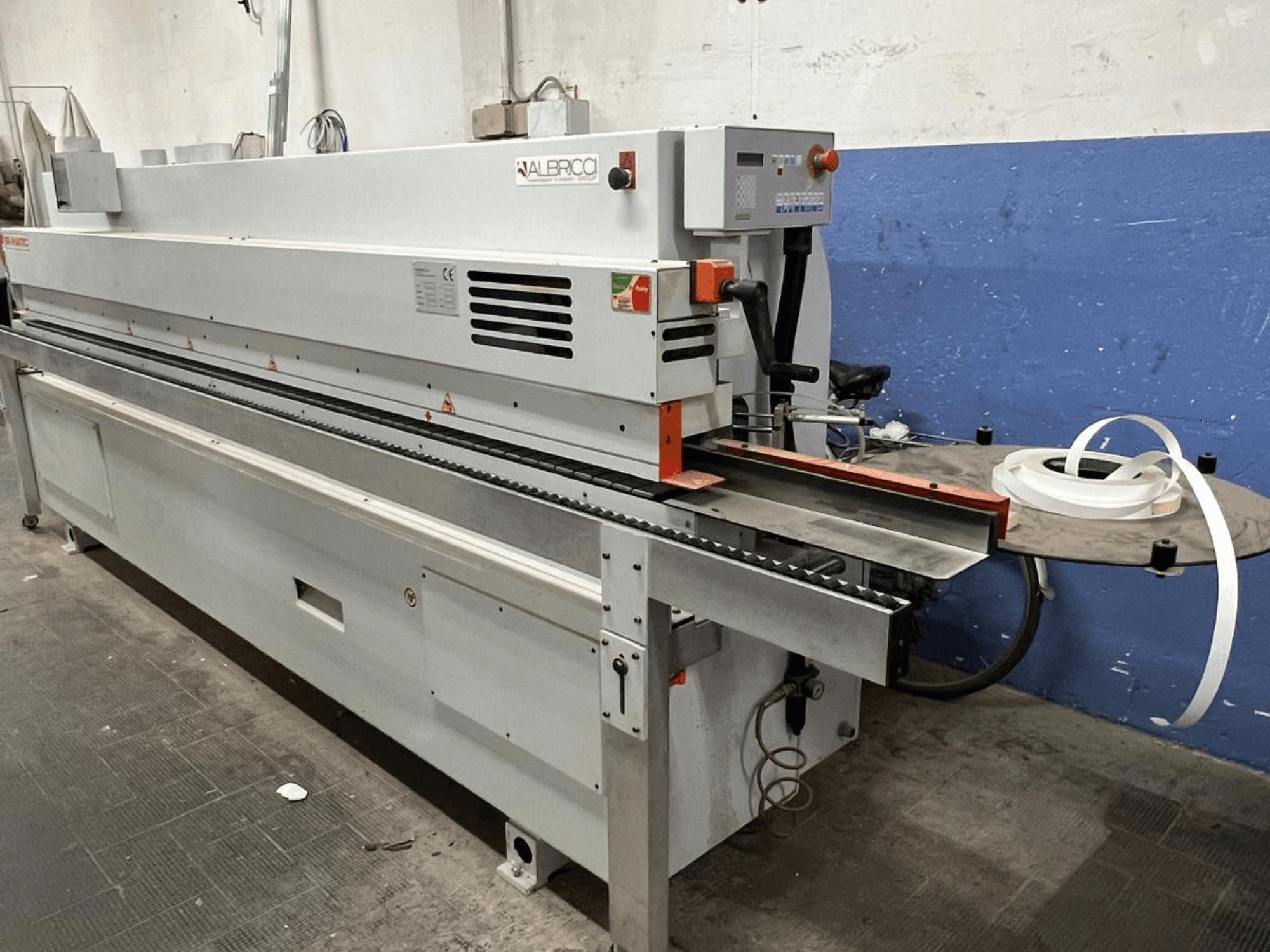 ALBRICO kraštų juostų klijavimo mašinos vaizdas iš priekio su ilga konvejerio juosta ir pritvirtintu lipnios juostos ritiniu.