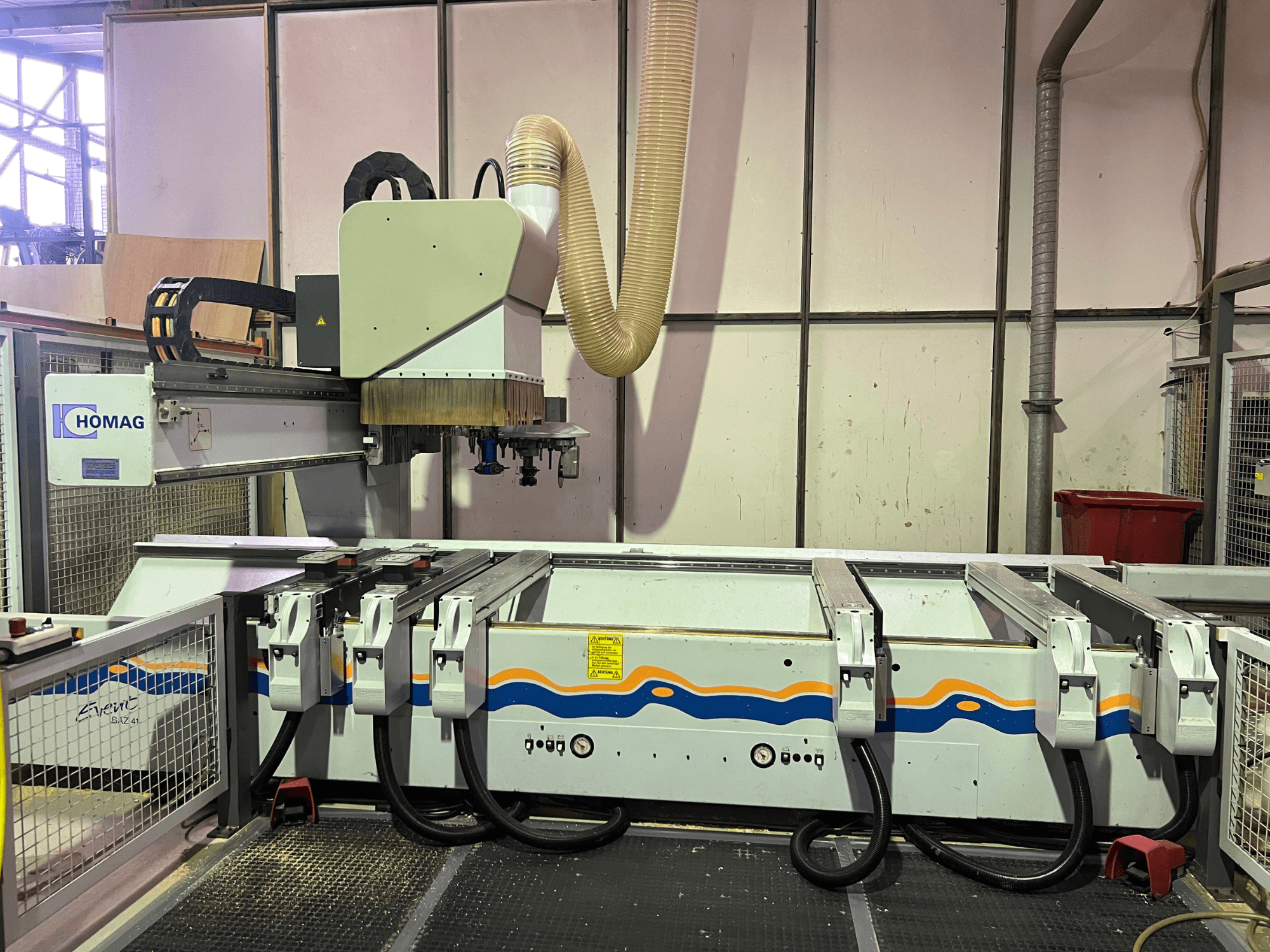 HOMAG BIZ 4 CNC staklės dirbtuvėse; vaizdas iš priekio, kuriame matoma dulkių ištraukimo sistema ir valdymo skydelis su kabeliais.