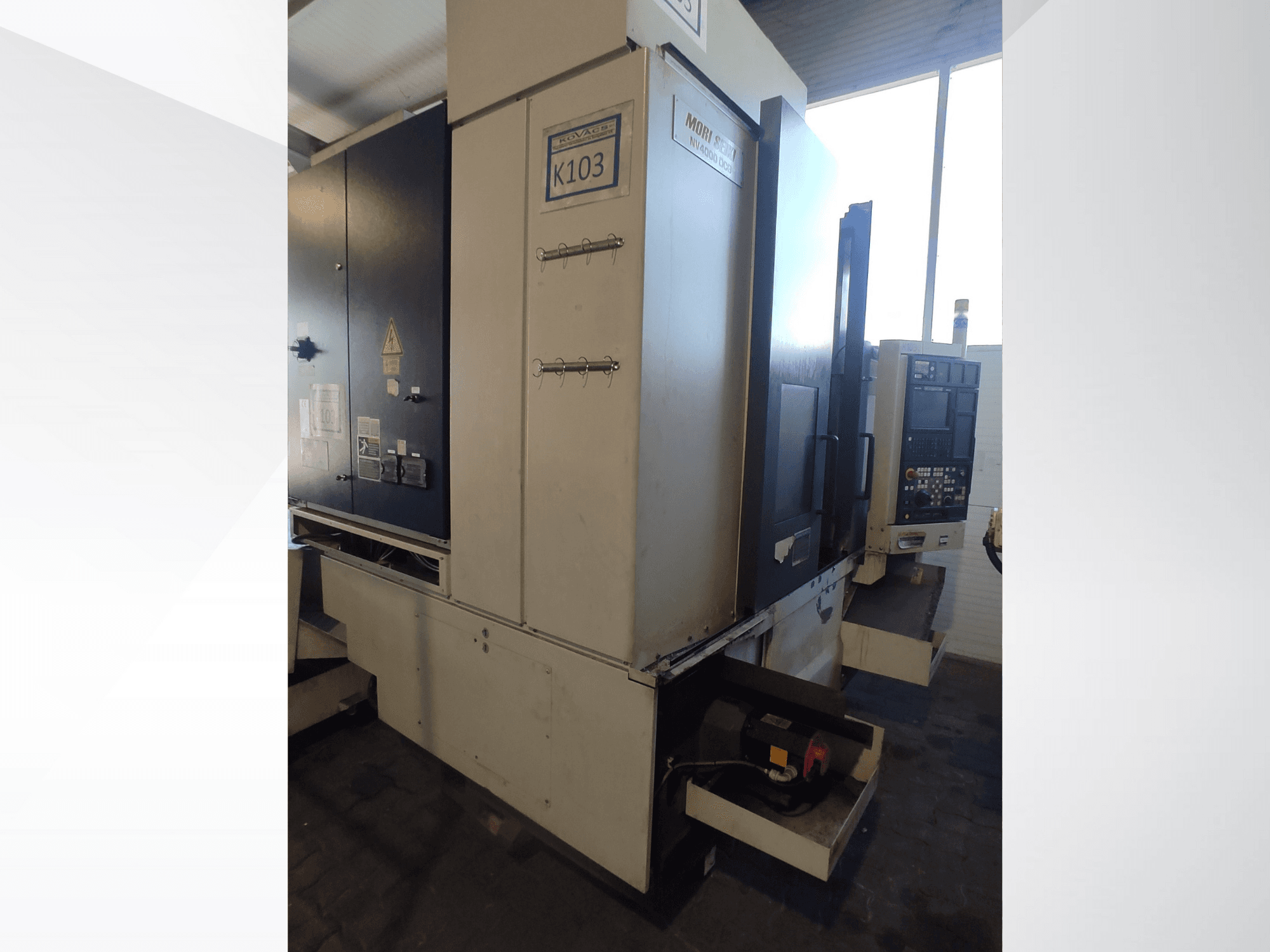 Mori Seiki K103 CNC staklės iš priekio, matomas valdymo skydelis, saugos etiketės ir elektros komponentai.