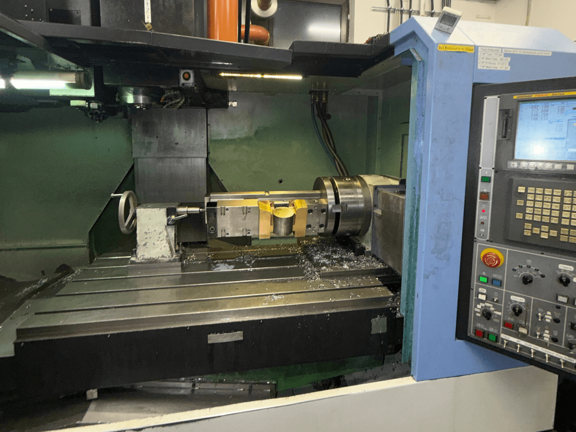Vertikalusis apdirbimo centras „DOOSAN DNM650“, vaizdas iš priekio, kuriame matomas pjovimo įrankis ir valdymo pultas, o ant stalo matomos metalo drožlės.