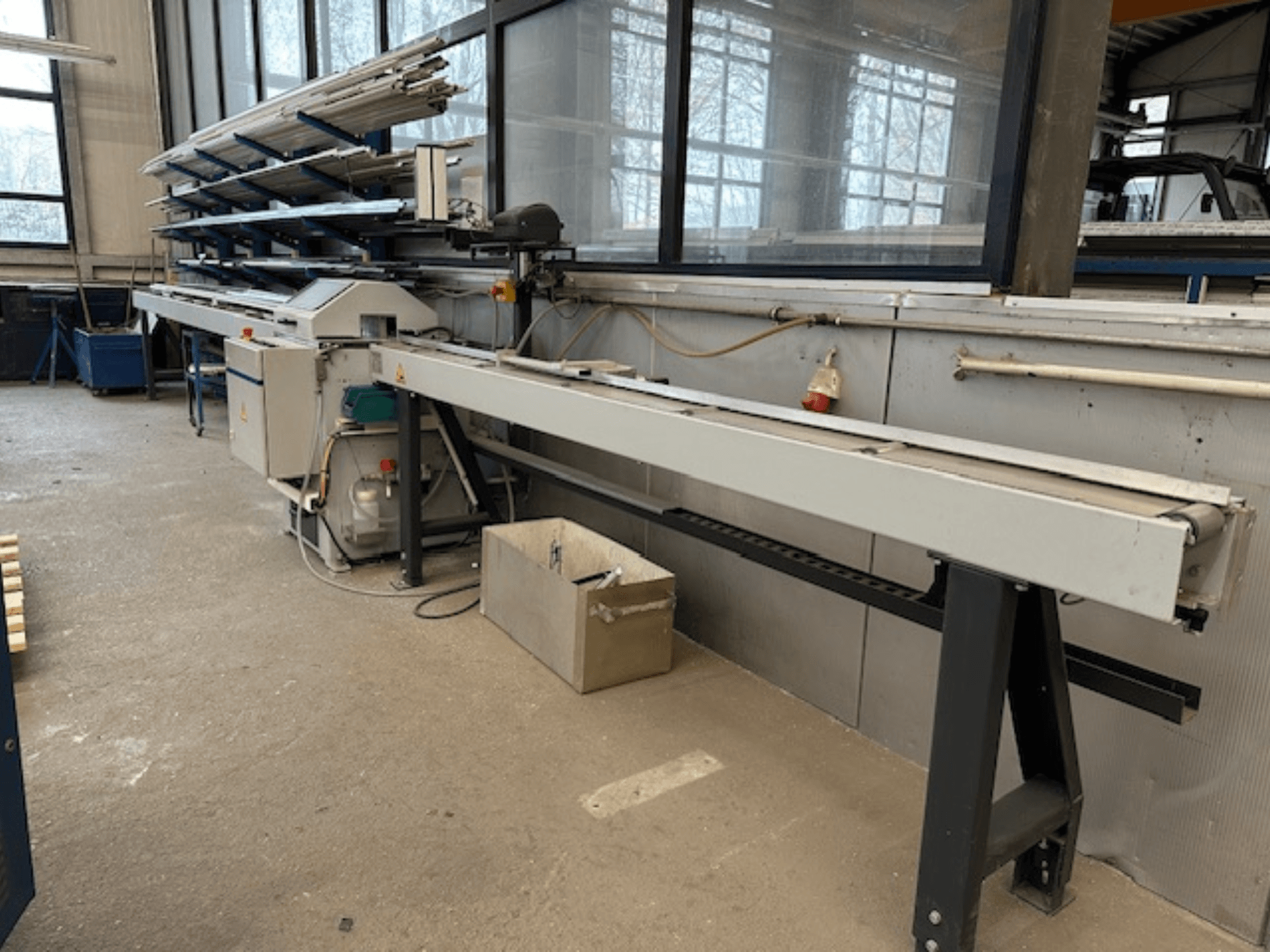 SCHÜCO PGS diskinis metalo pjūklas dirbtuvėse, šoninis vaizdas su reguliuojama pjovimo lova ir virš jos esančia metalinių profilių lentyna.