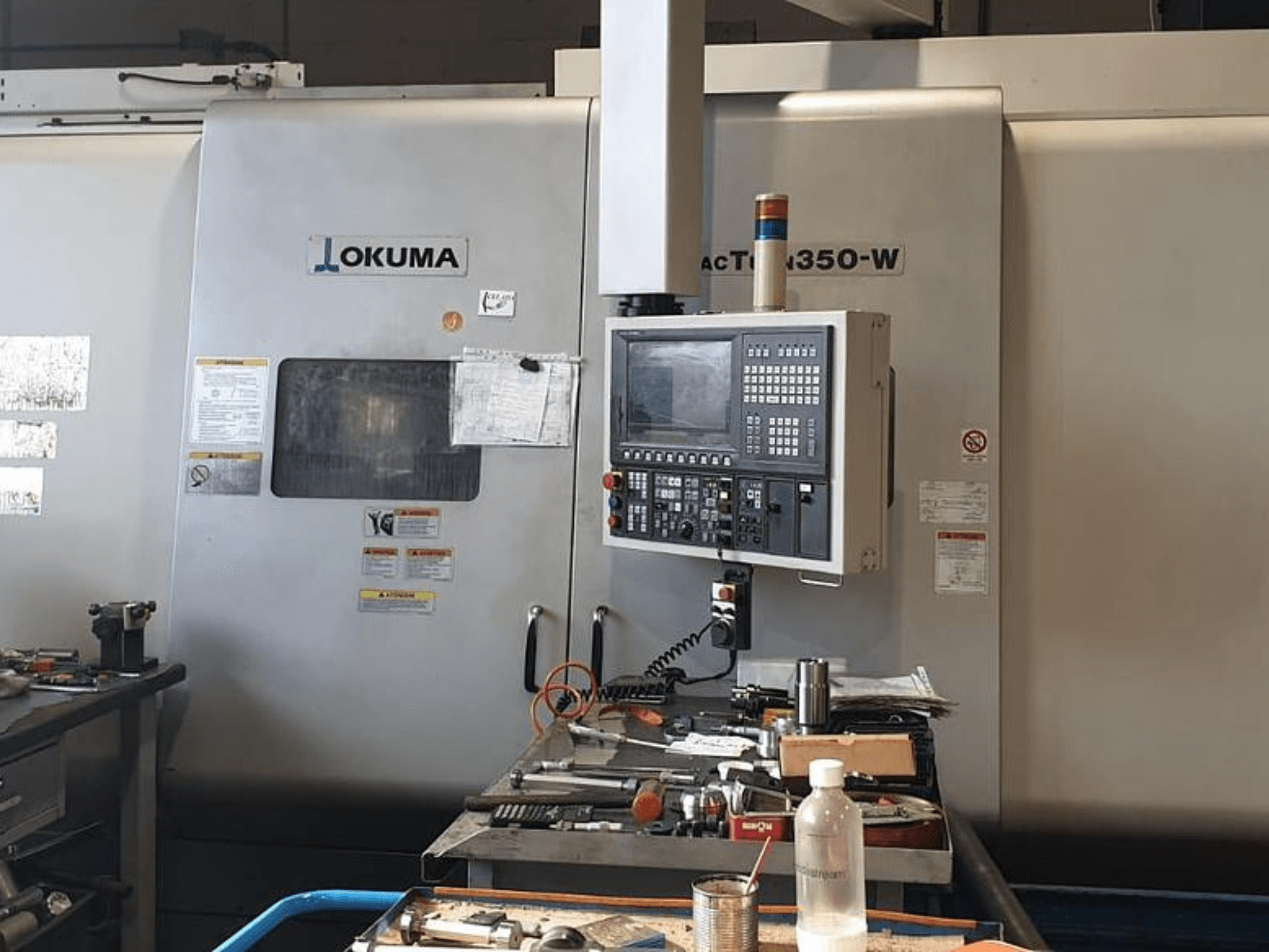 Okuma MAC TURN 350-W tekinimo frezavimo centras, vaizdas iš priekio su valdymo pultu ir darbo vieta, užgriozdinta įrankiais ir buteliu.