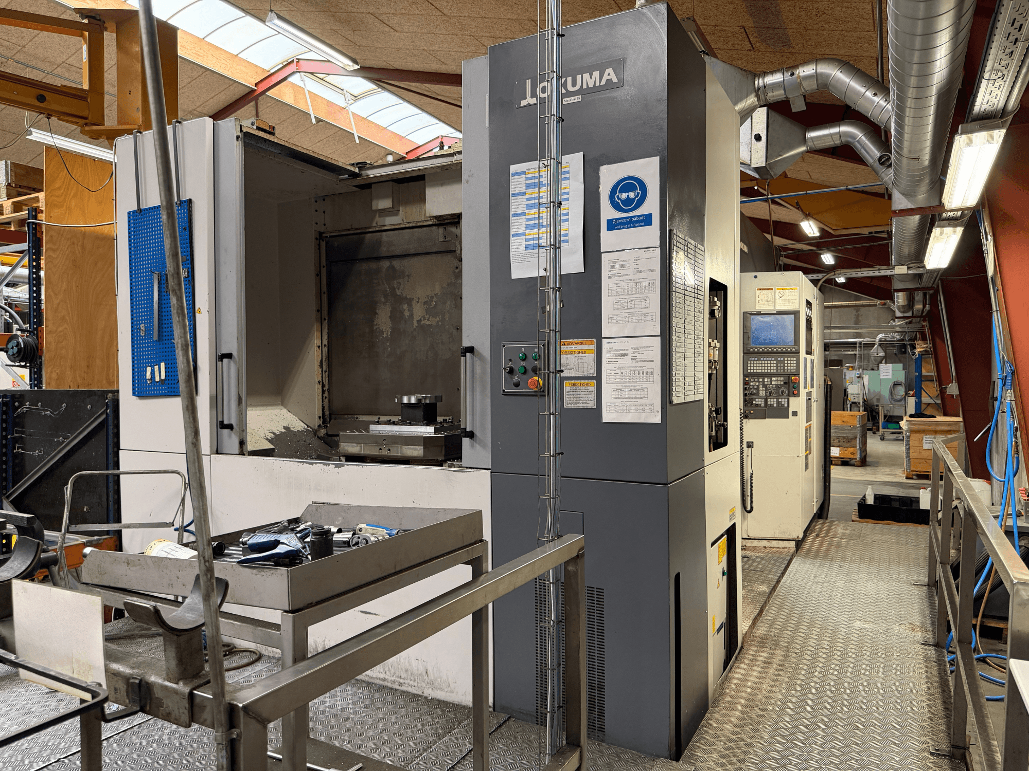 „Okuma MA-600HB“ horizontalus apdirbimo centras, vaizdas iš priekio, kuriame matomi ant metalinio stalo esantys įrankiai ir staklių valdymo elementai.