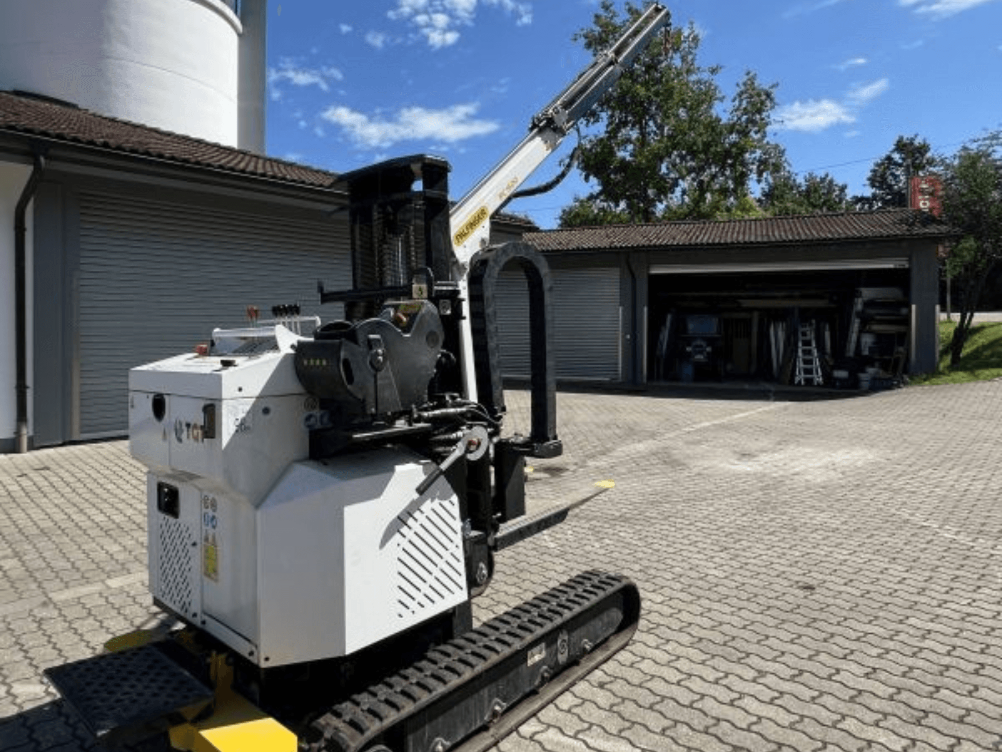 TGI statybinis mini kranas, šoninis vaizdas, matyti vikšrai ir sumontuota strėlė, pastatytas ant mūrinio paviršiaus prie garažo.