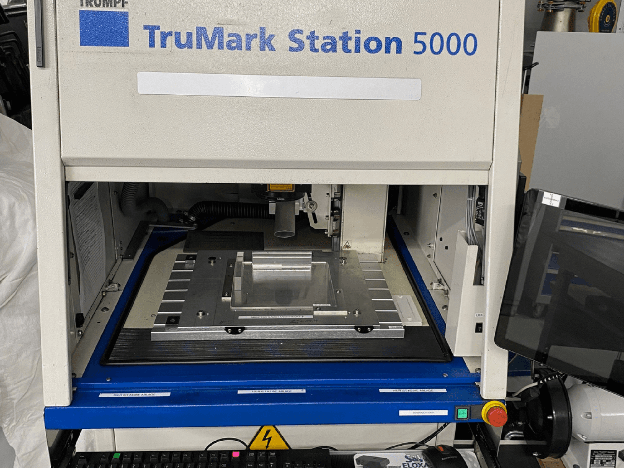 "TruMark Station 5000" lazerinio žymėjimo mašinos priekinis vaizdas, kuriame matomas darbo stalas, valdikliai ir stebėjimo ekranas.