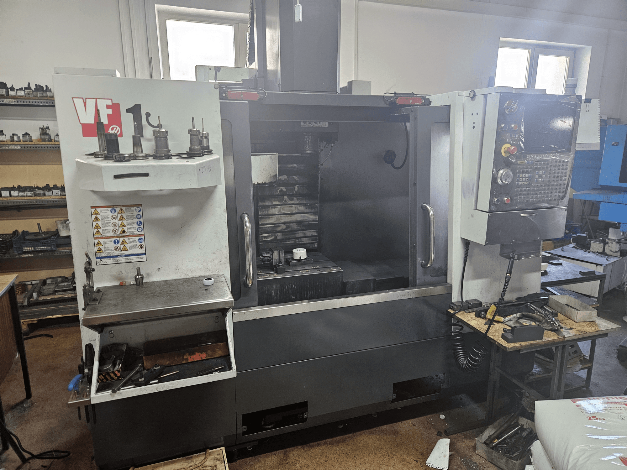 HAAS VF-1 vertikalusis apdirbimo centras, vaizdas iš priekio su įrankiais šone, matomas valdymo skydelis ir darbo vieta fone.