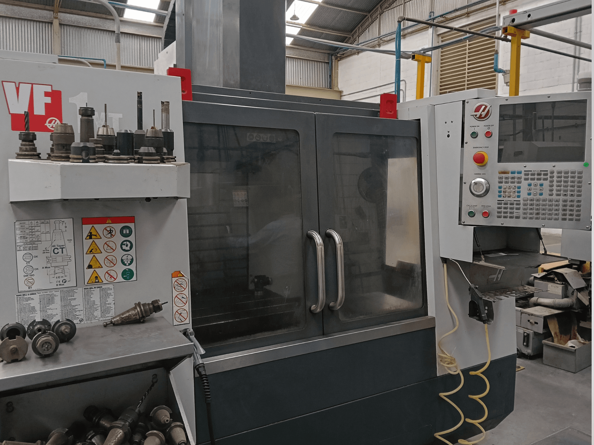 HAAS VF-1YT-3 vertikalaus apdirbimo centro vaizdas iš priekio, kuriame matomi valdymo elementai, įrankių laikikliai ir saugos etiketės.