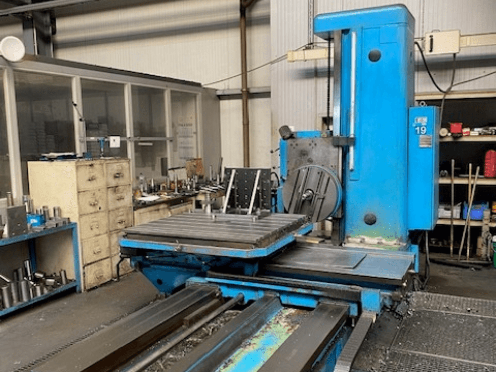 Mėlynos spalvos frezavimo staklės, 19 modelio, iš priekio, ant dirbtuvių grindų, fone - įrankiai ir sandėliavimo spinta.