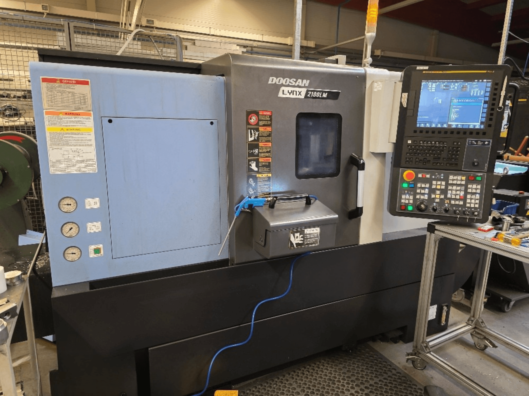 CNC staklės "Doosan Lynx 2100LM CNC", vaizdas iš priekio, su valdymo skydeliu, manometrais ir saugos įspėjimais.