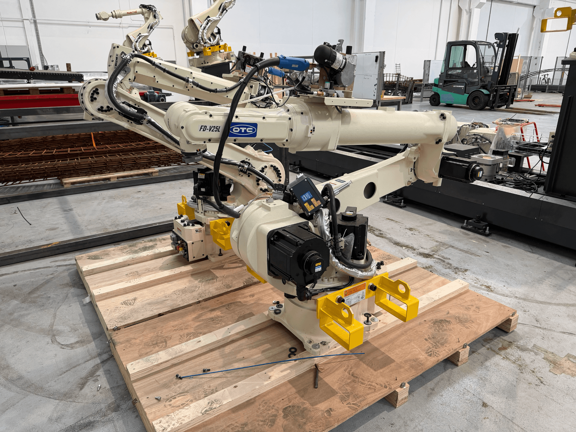 „OTC Daihen FD19-V25L“ robotinė ranka, vaizdas iš priekio, su geltonomis griebtuvų letenomis ir matomais mechaniniais komponentais ant medinės paletės.