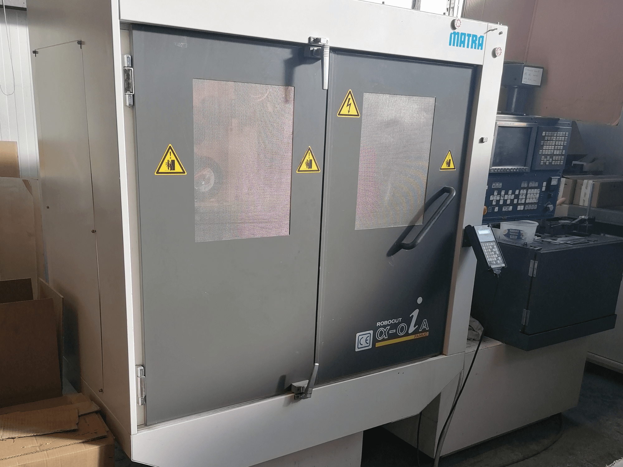 "Matra" "Robocut α-oZα" staklės, priekinis vaizdas, kuriame matomi saugos įspėjimai, valdymo skydelis ir dvejos durys su tinkliniais langais.