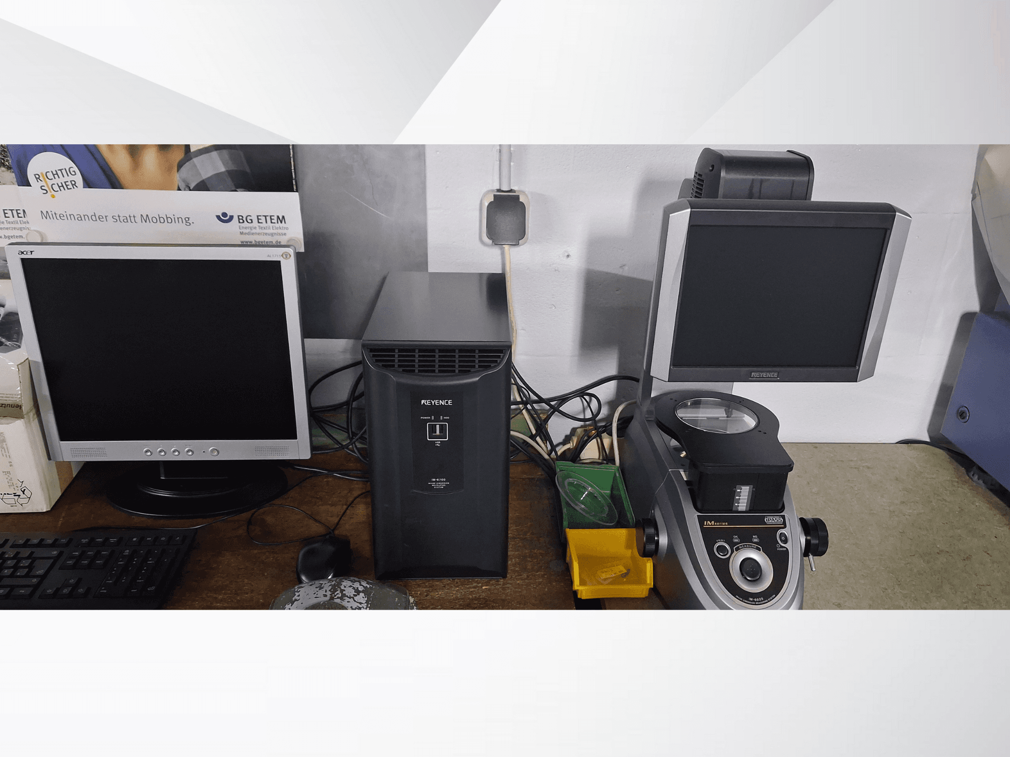 Du kompiuterių monitoriai, vienas "Acer", kitas "Keyence", šalia juodo prietaiso ir mikroskopo, žiūrint iš priekio.