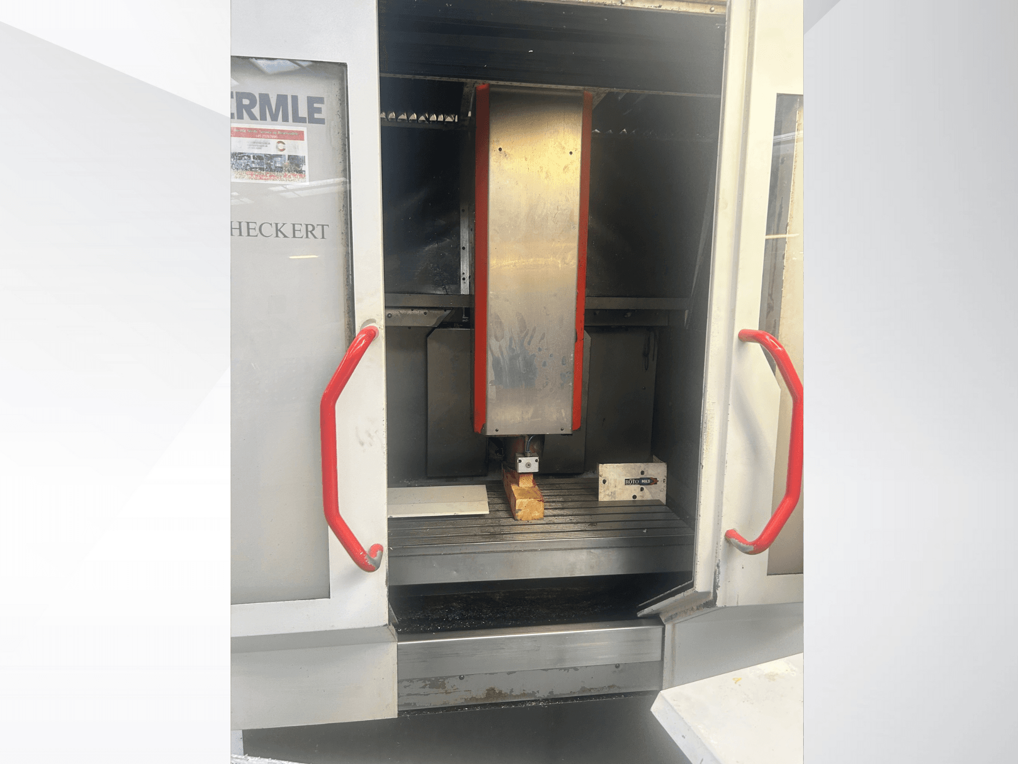 "Hermle" CNC staklių vaizdas iš priekio - metalinis vidus, raudonos rankenos ir ant stalo esantis ruošinys.