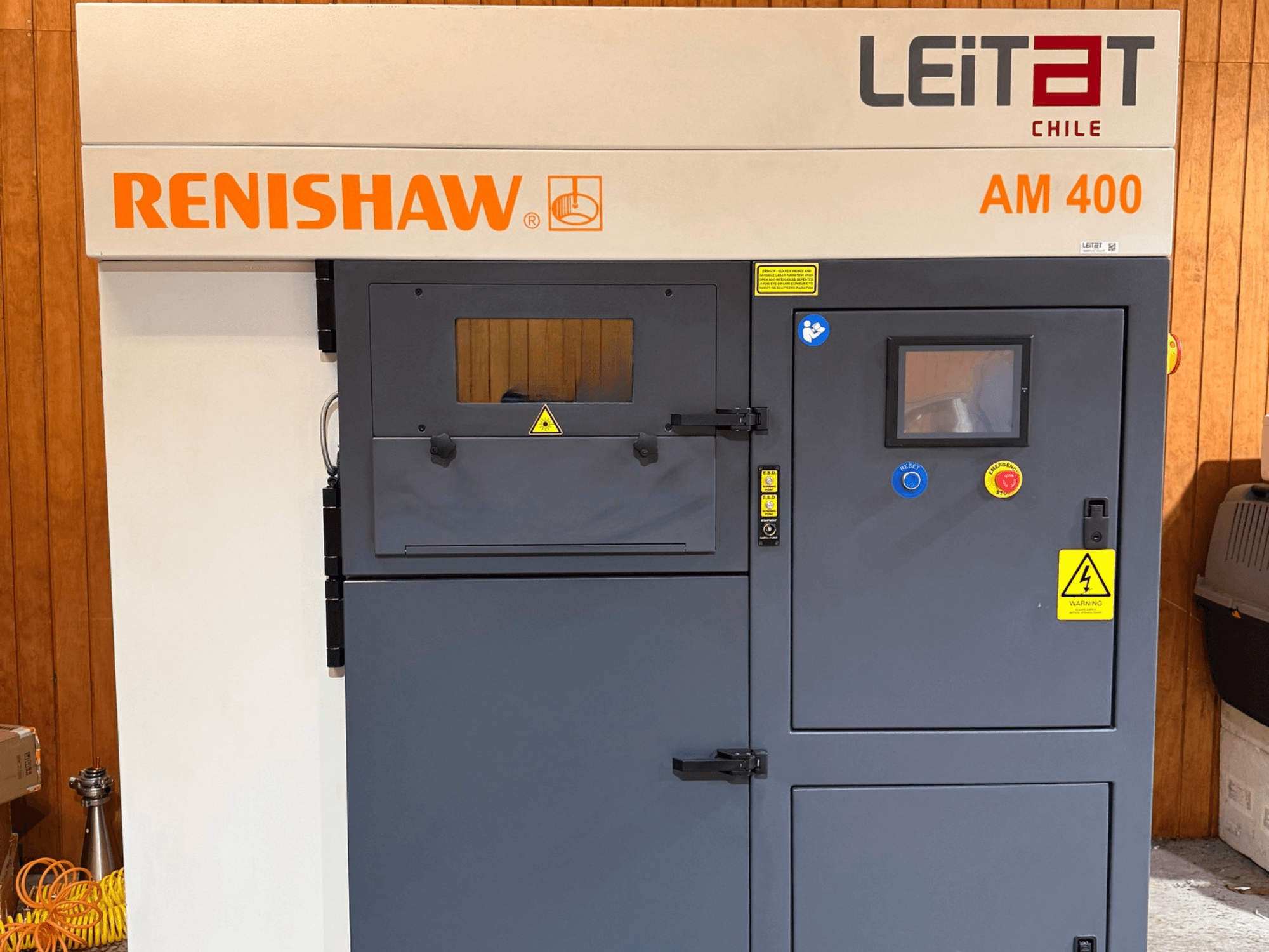 „RENISHAW AM400“ metalo 3D spausdintuvo vaizdas iš priekio, kuriame matomas valdymo skydelis, įspėjamieji užrašai ir prieiga prie techninės priežiūros vietų.